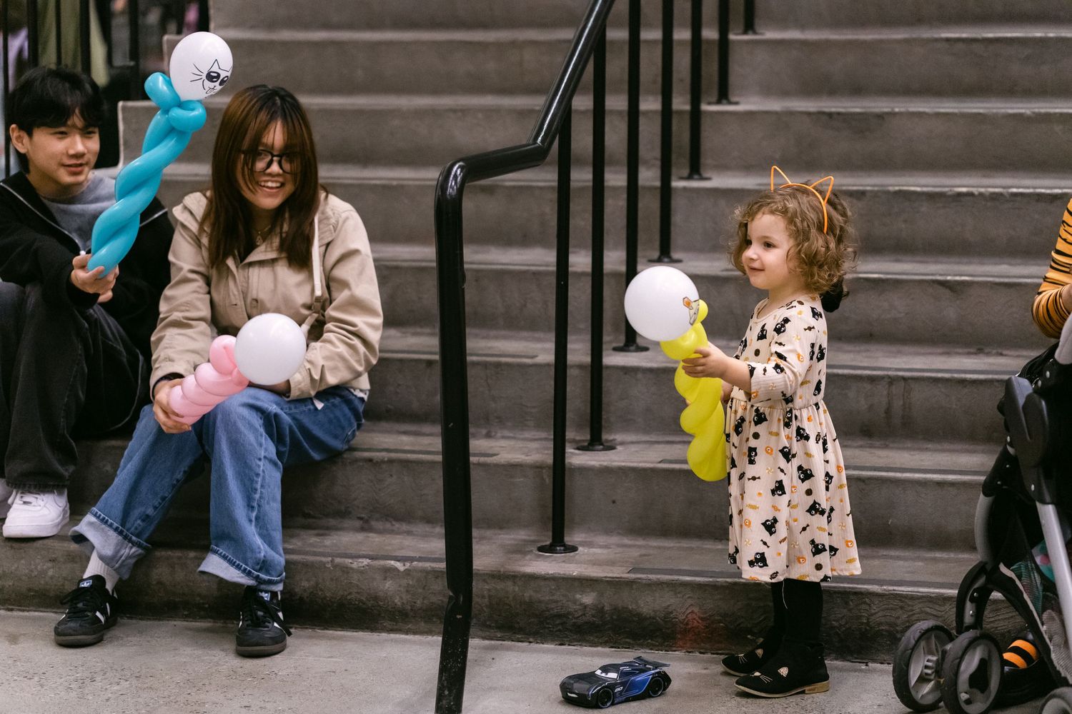A toddler girl holds balloons near stairs while a woman and child sit on steps at an indoor event.