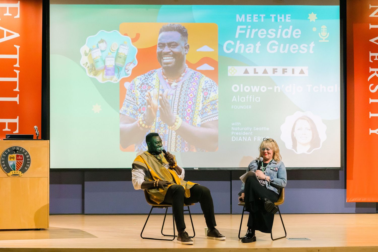 Fireside chat event at a university with two speakers seated on stage before a large presentation screen.