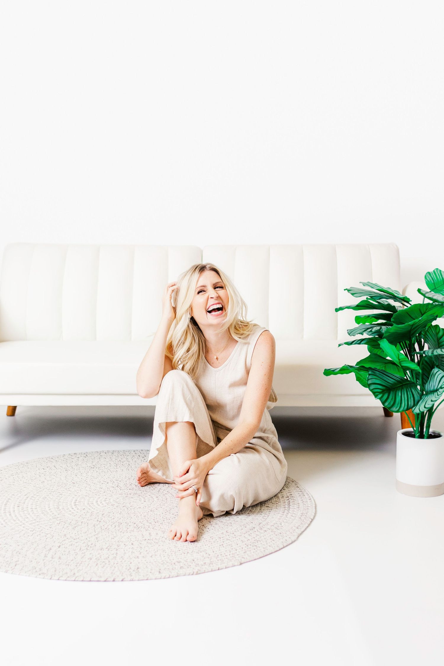 Laughing blonde woman sitting barefoot on a round rug beside a white sofa and green plant in a bright room.