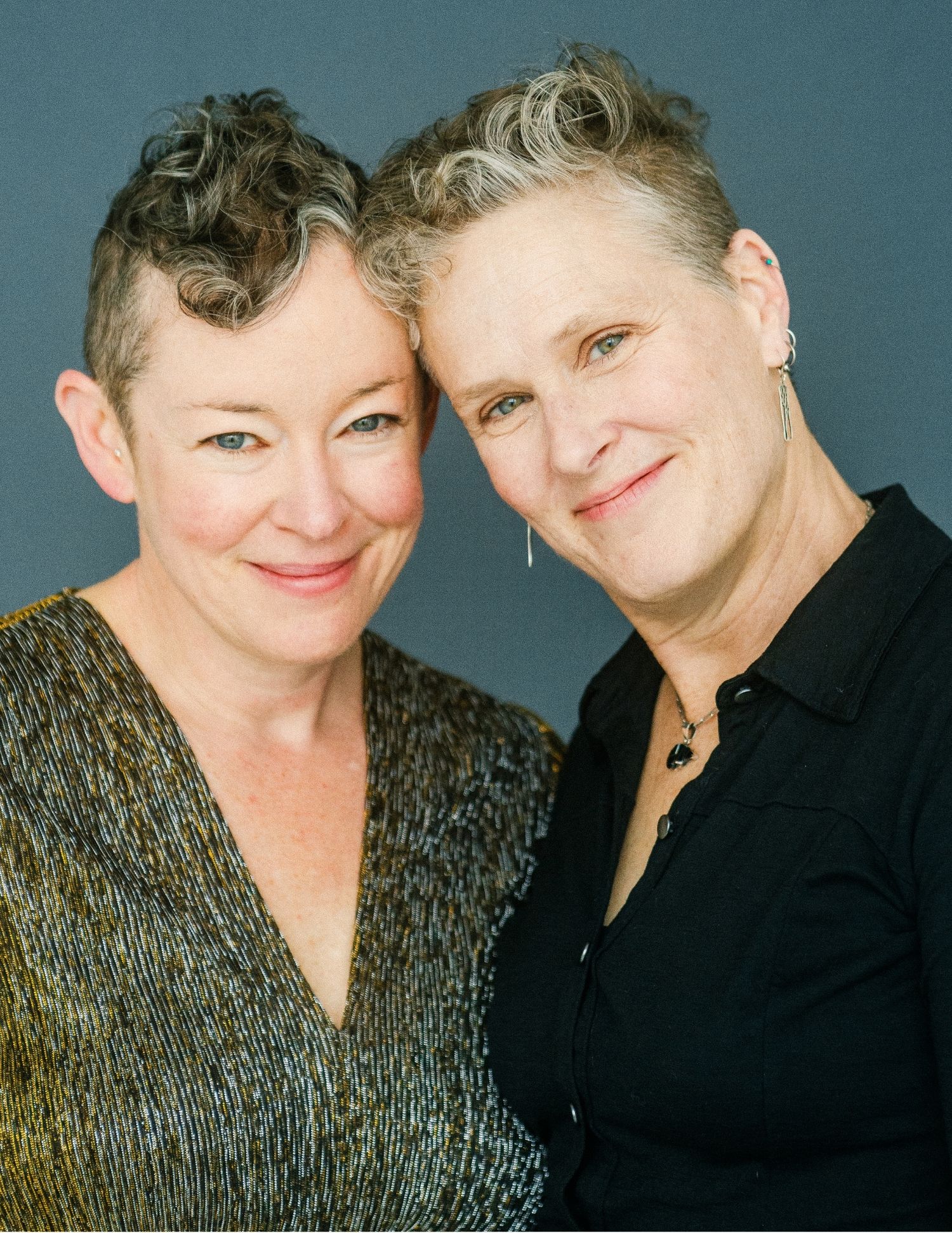Two smiling middle-aged women with short gray hair pose cheek to cheek against a gray studio background.