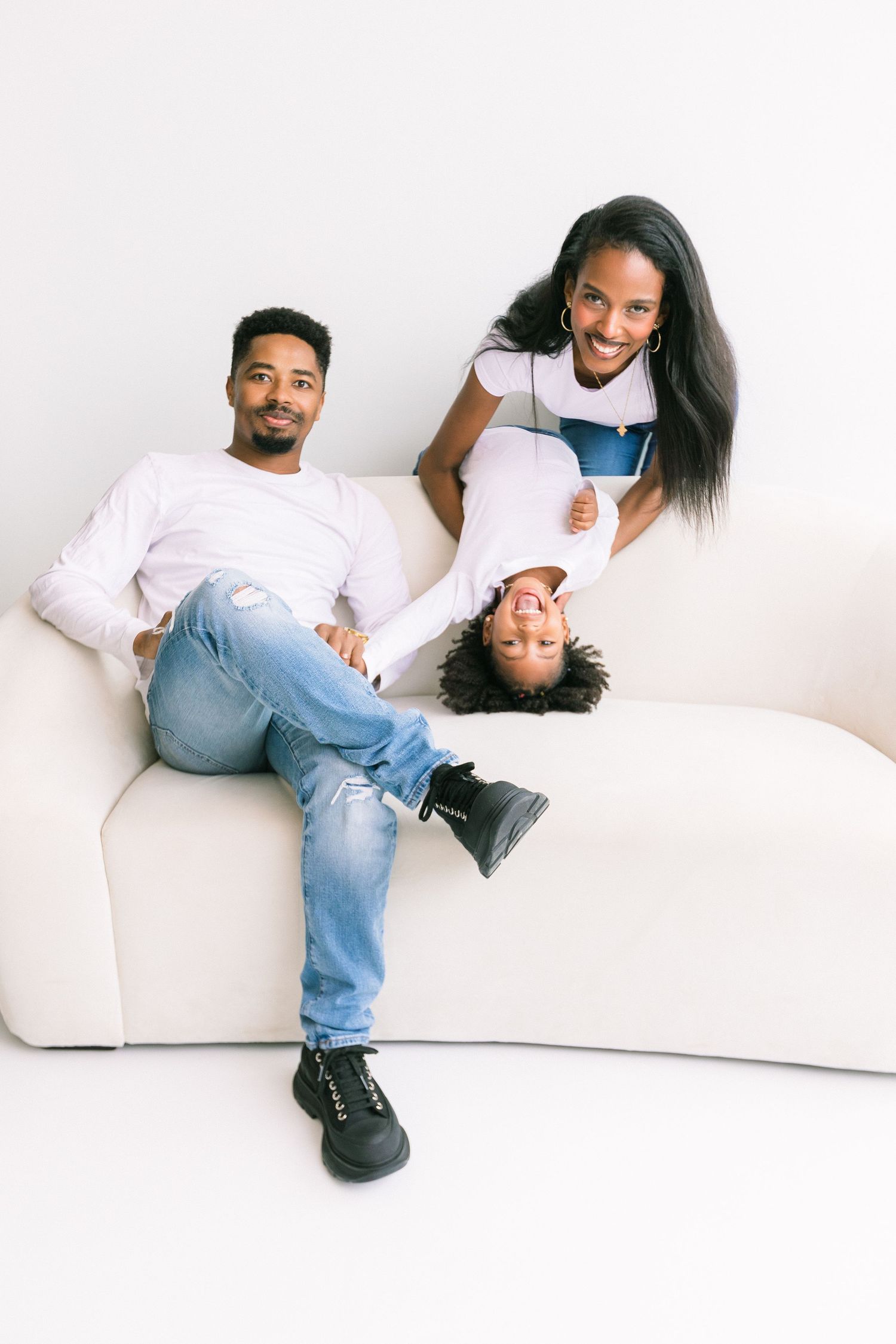 Happy Black family of three relaxing on a white couch, with child playfully hanging upside down.