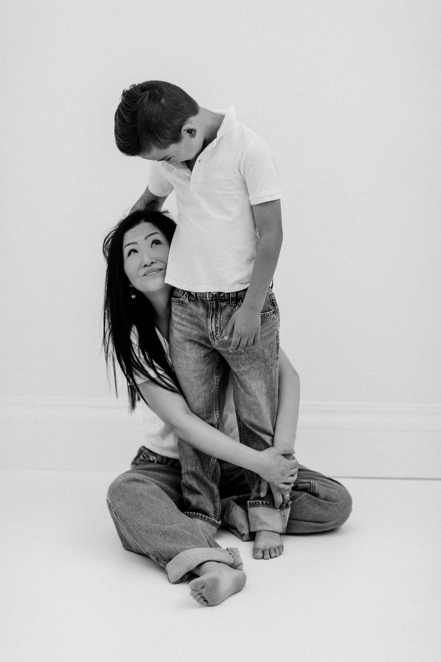 Black and white photo of a woman sitting on floor hugging a boy standing behind her in matching jeans and white tops.