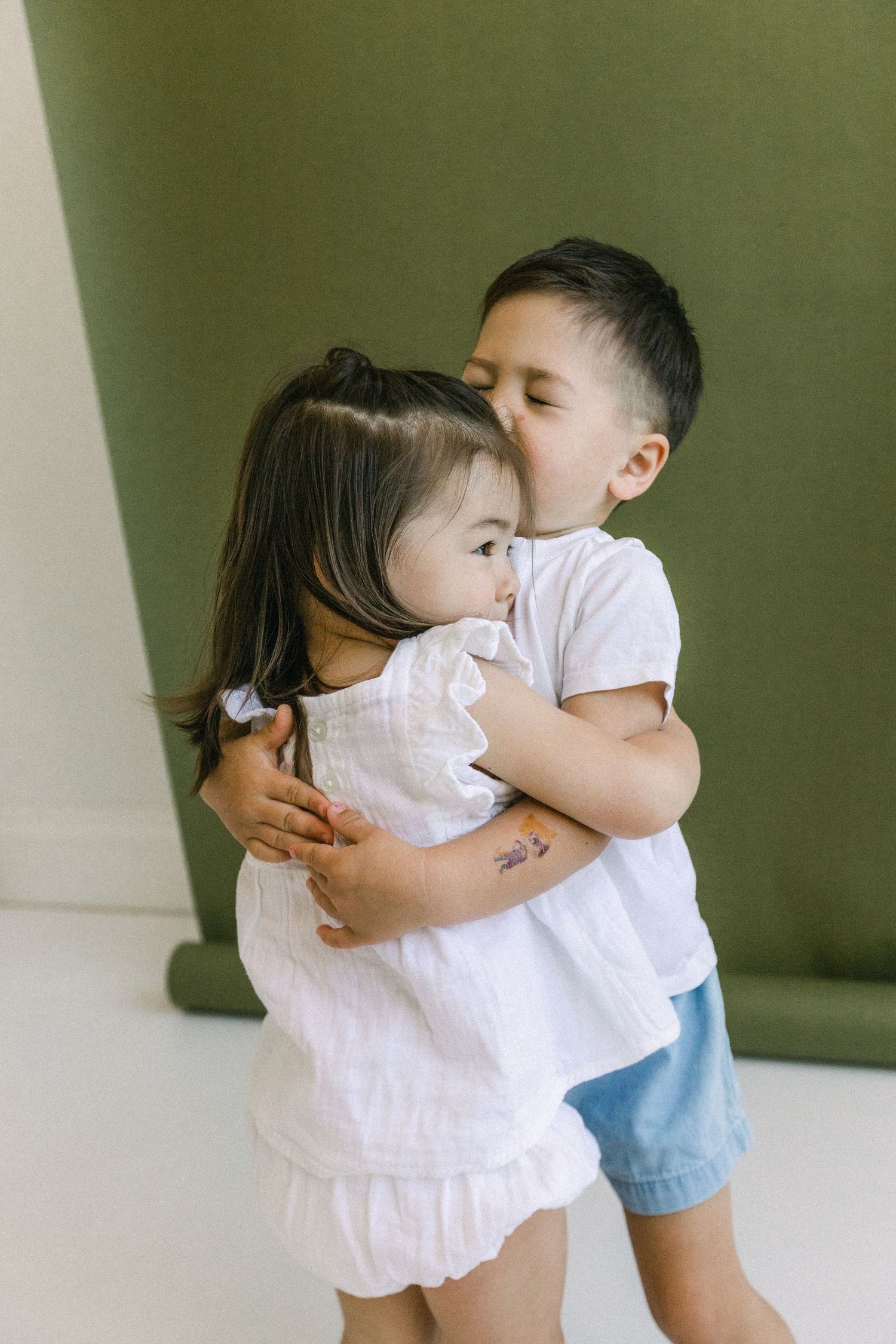 Two young siblings share a sweet hug and kiss, dressed in white tops against an olive green background.