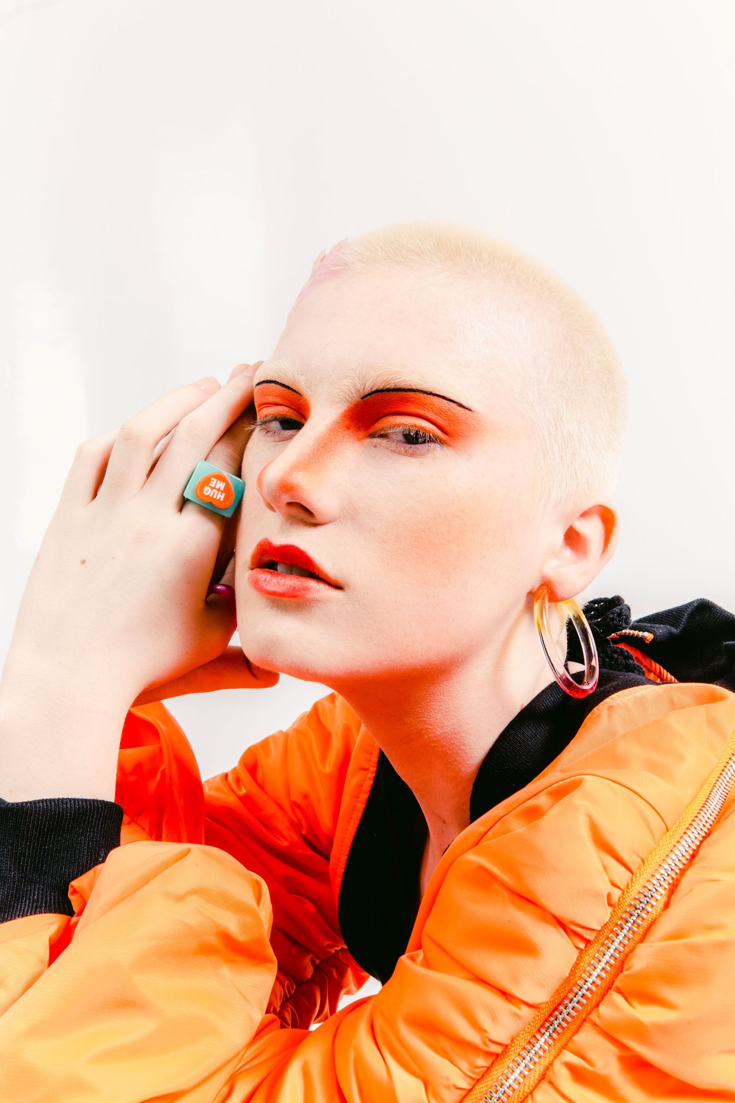 Bold fashion portrait of a model with shaved head wearing orange makeup and orange puffer jacket.