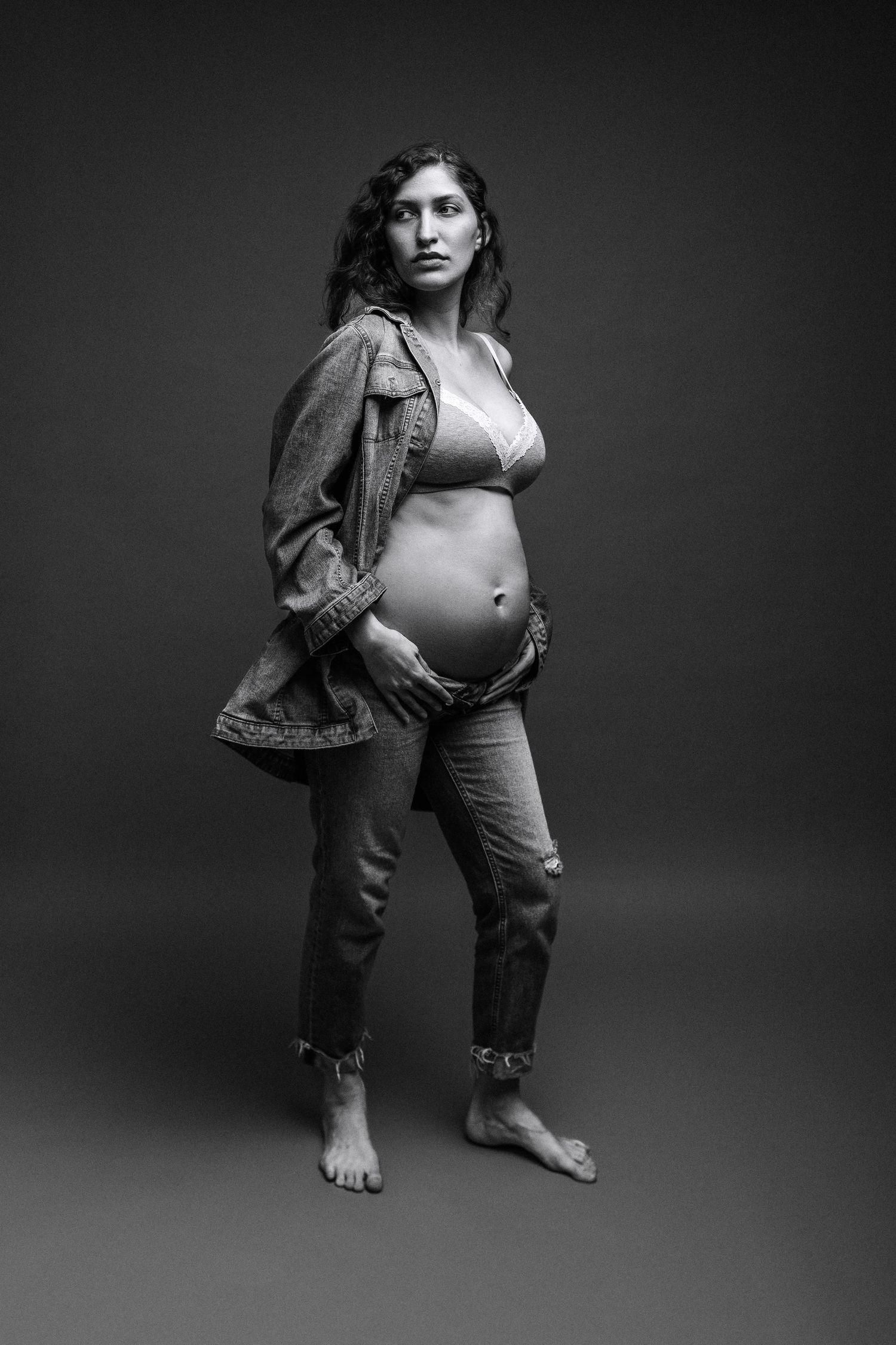 Black and white portrait of a pregnant woman in denim jacket and jeans, cradling her baby bump in a studio.