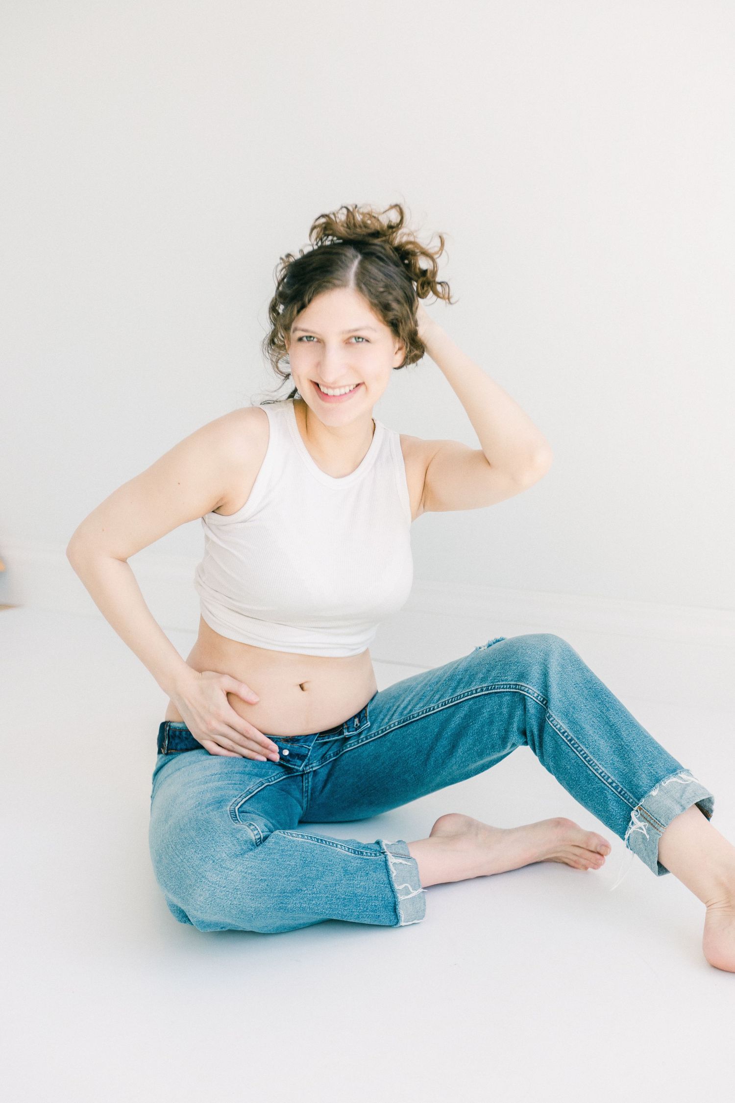 Smiling young woman in white crop top and blue jeans sitting on floor against white background.