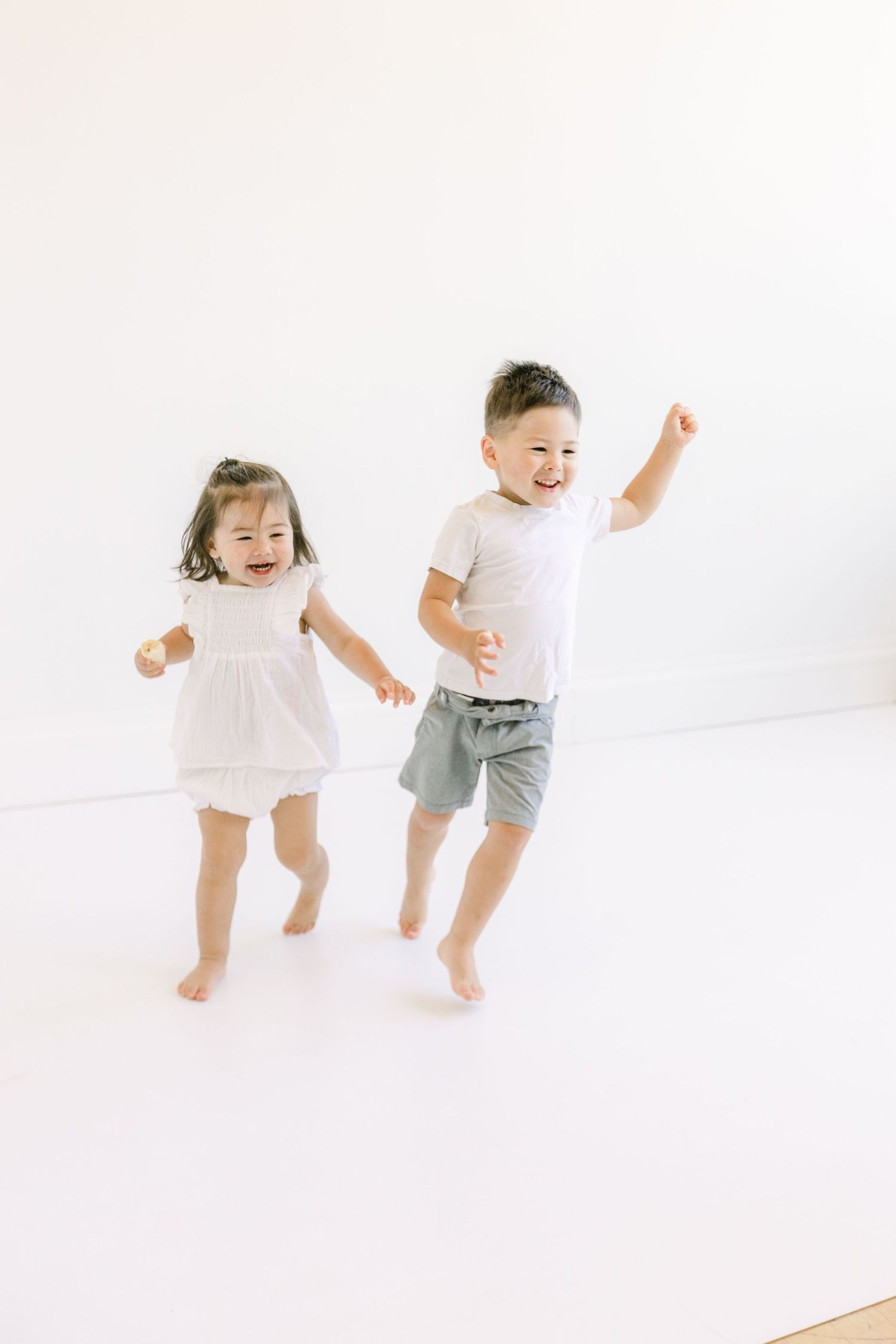 Two young children laughing and jumping joyfully in a bright white studio, wearing casual light-colored outfits.