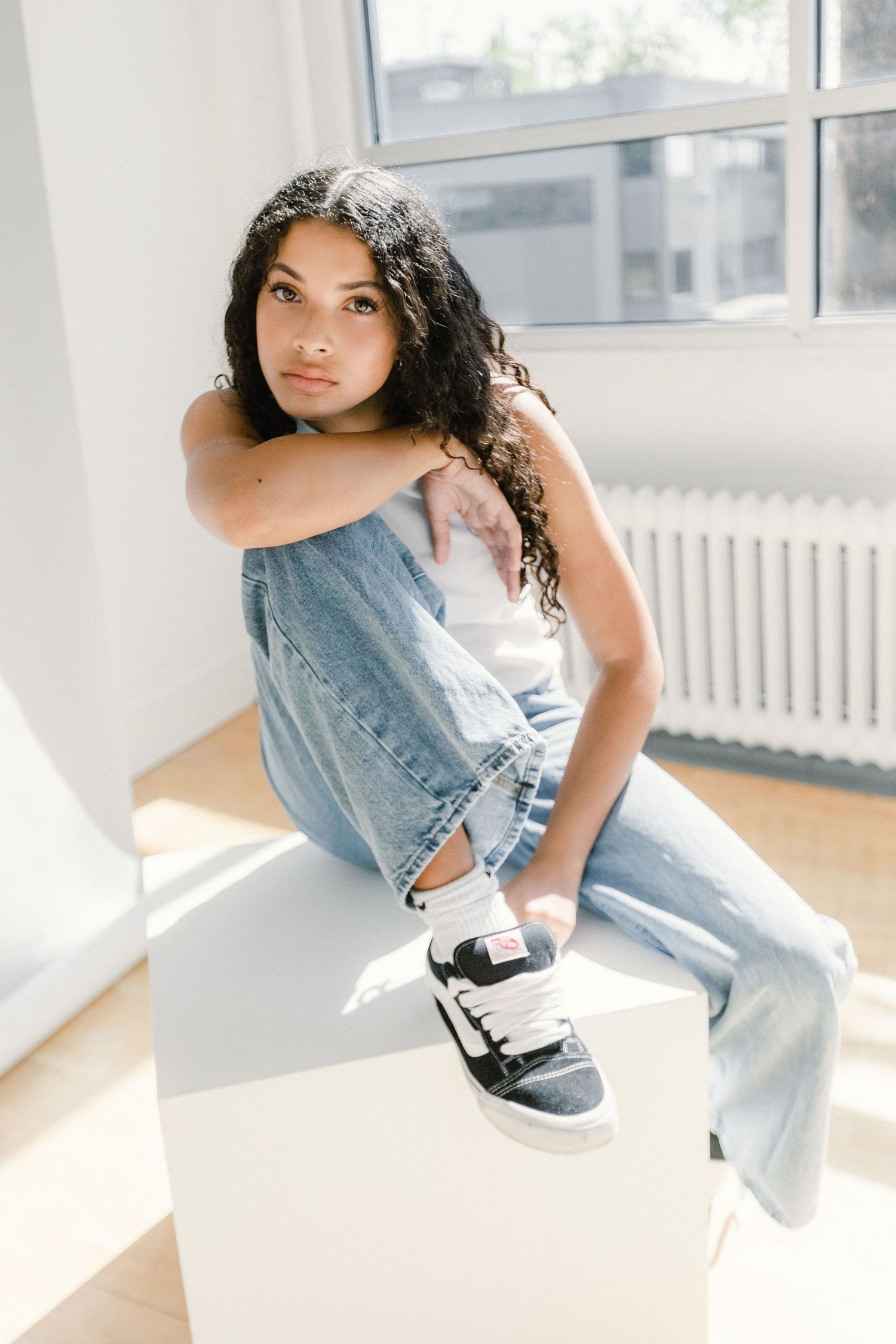 A young woman with curly hair sitting on a white cube in a sunlit studio wearing jeans and sneakers.