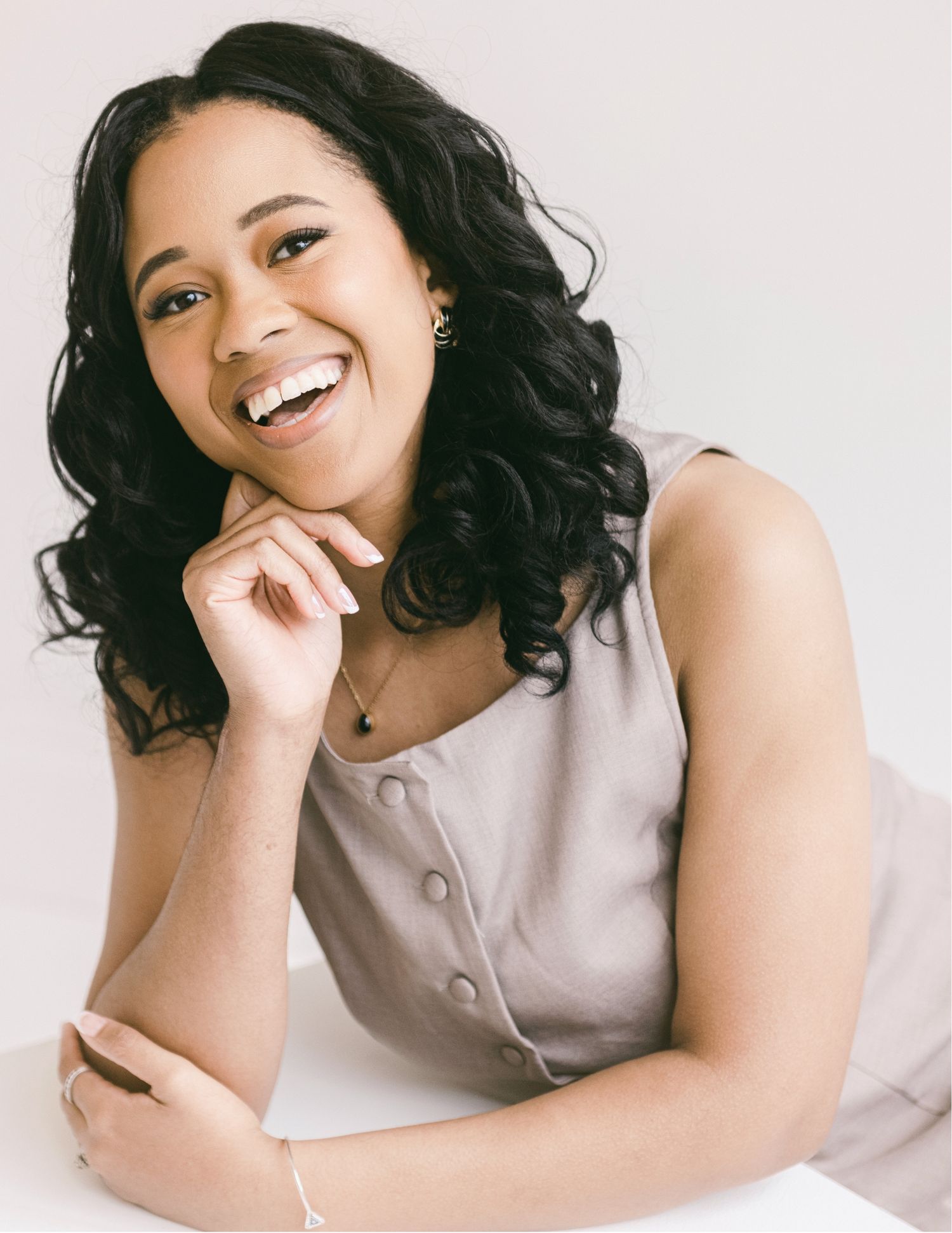 Smiling young woman with curly black hair wearing a sleeveless beige dress, posing confidently against a white background.