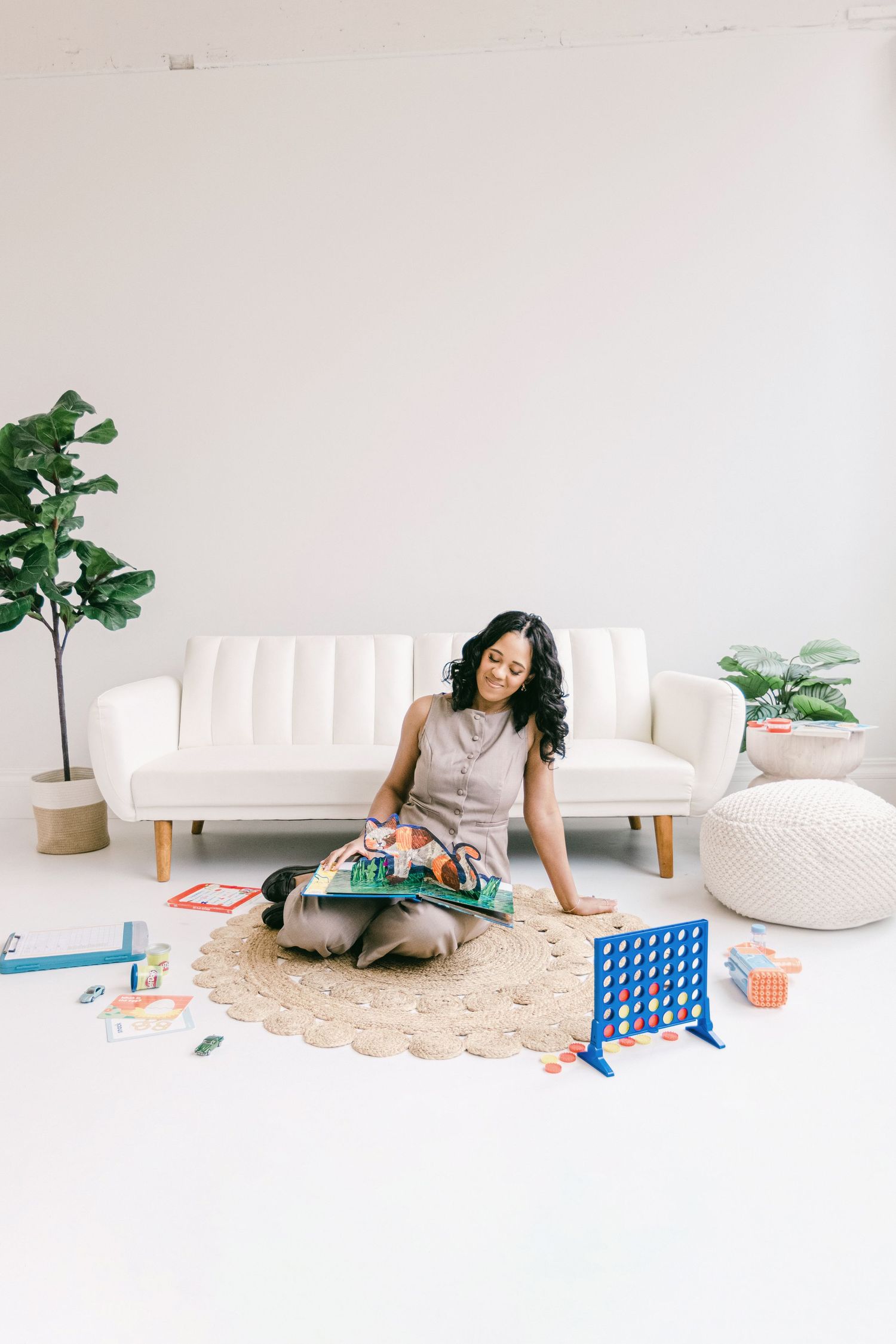 Woman sitting cross-legged on a jute rug reading a book surrounded by toys and games in a bright white minimalist room.