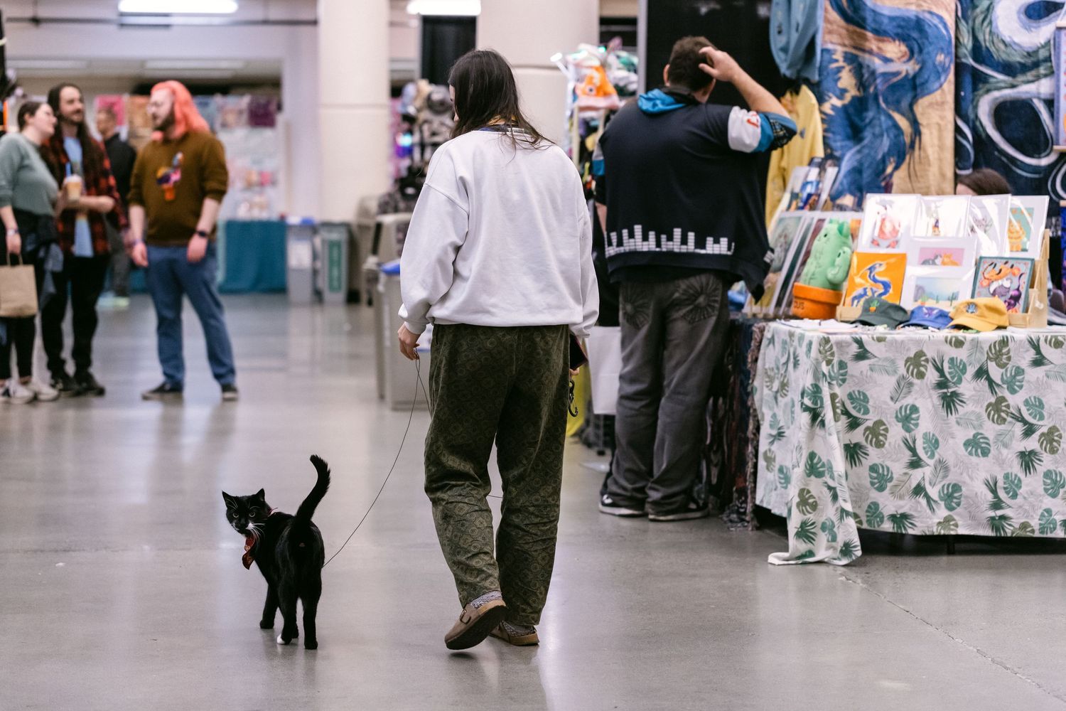 A person walks a black cat on a leash through a busy indoor art market with colorful displays.