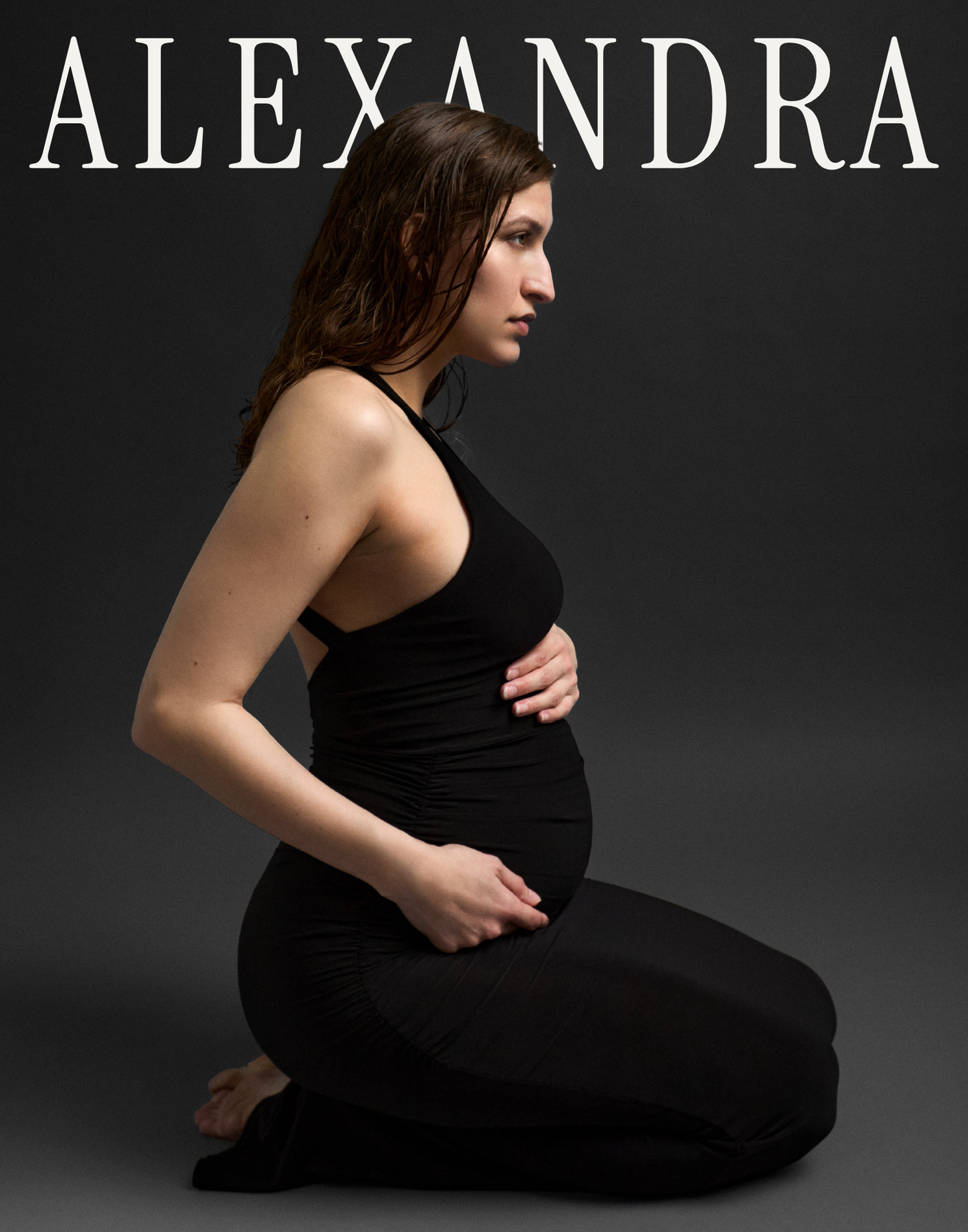 Pregnant woman in black outfit kneeling sideways, cradling baby bump, studio maternity portrait named Alexandra.