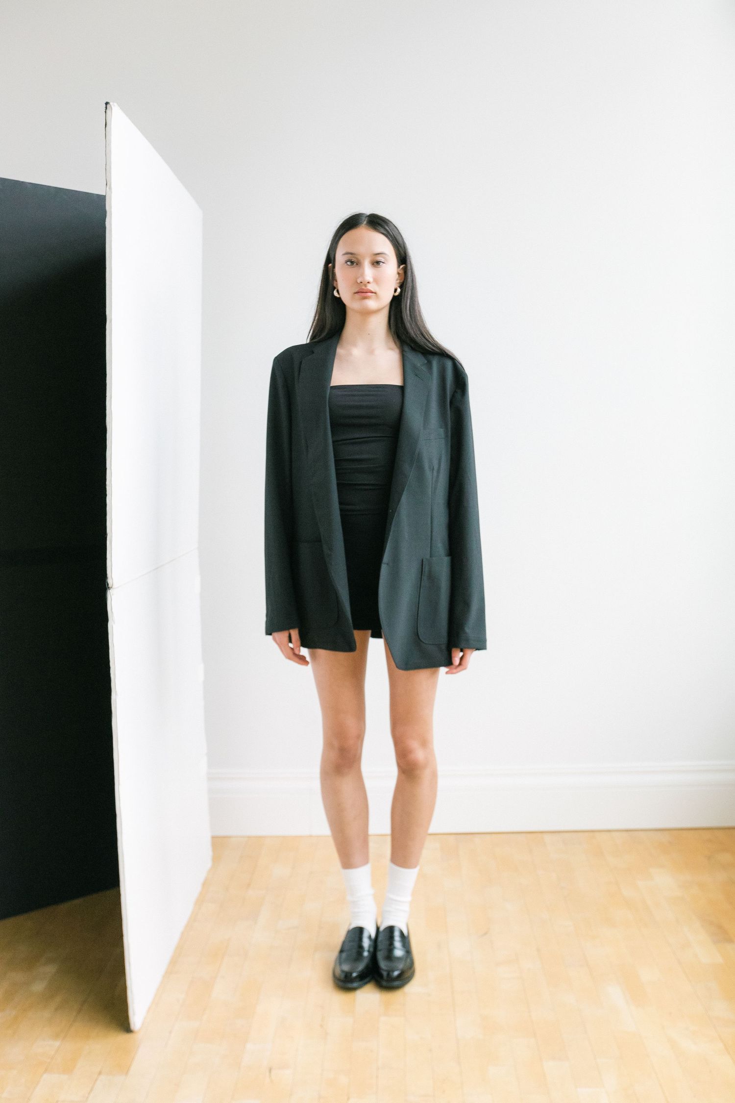 Woman in oversized black blazer, mini dress, white socks and loafers stands in minimalist studio setting.