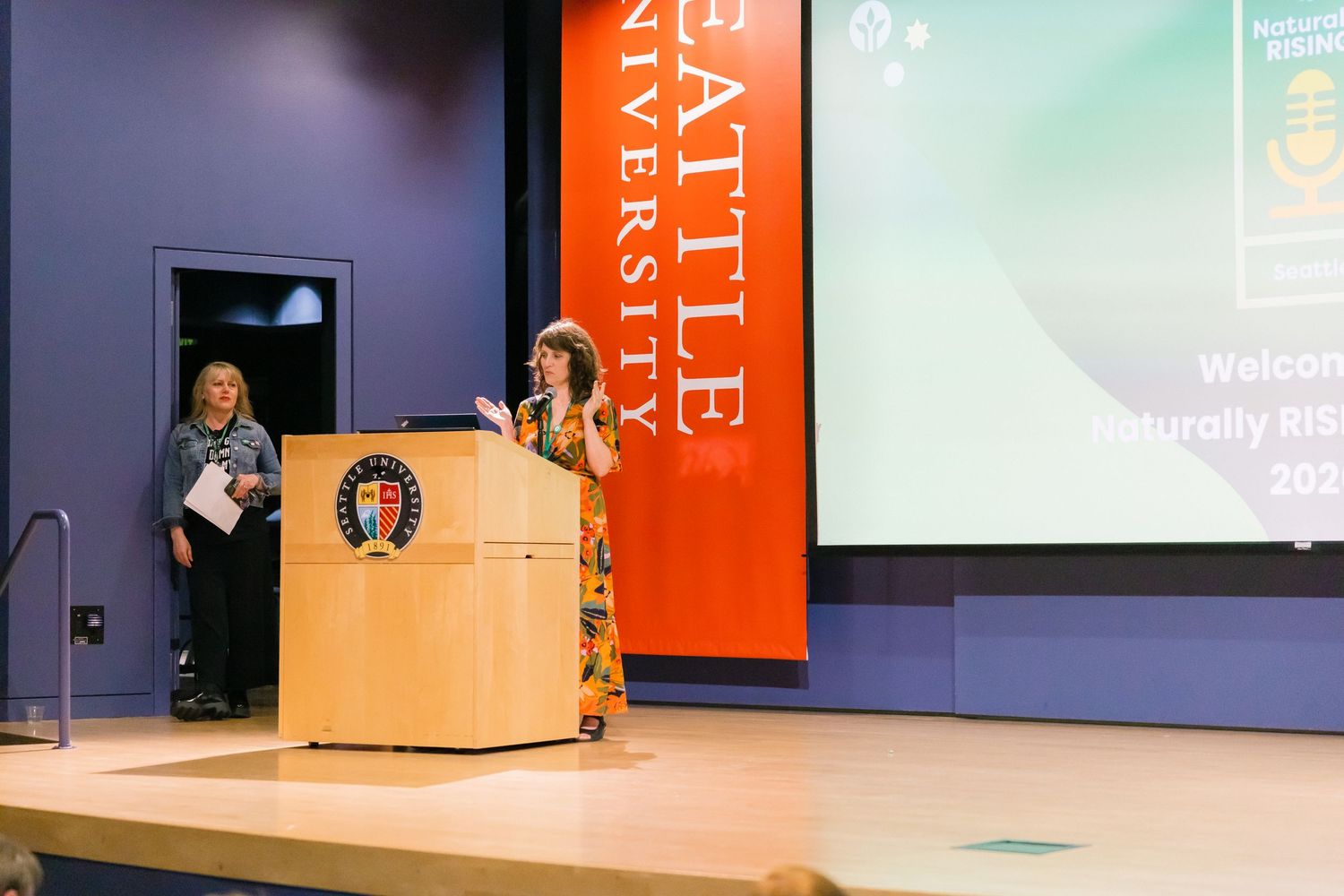 Woman speaking at podium on stage at Seattle University event with welcome screen backdrop.