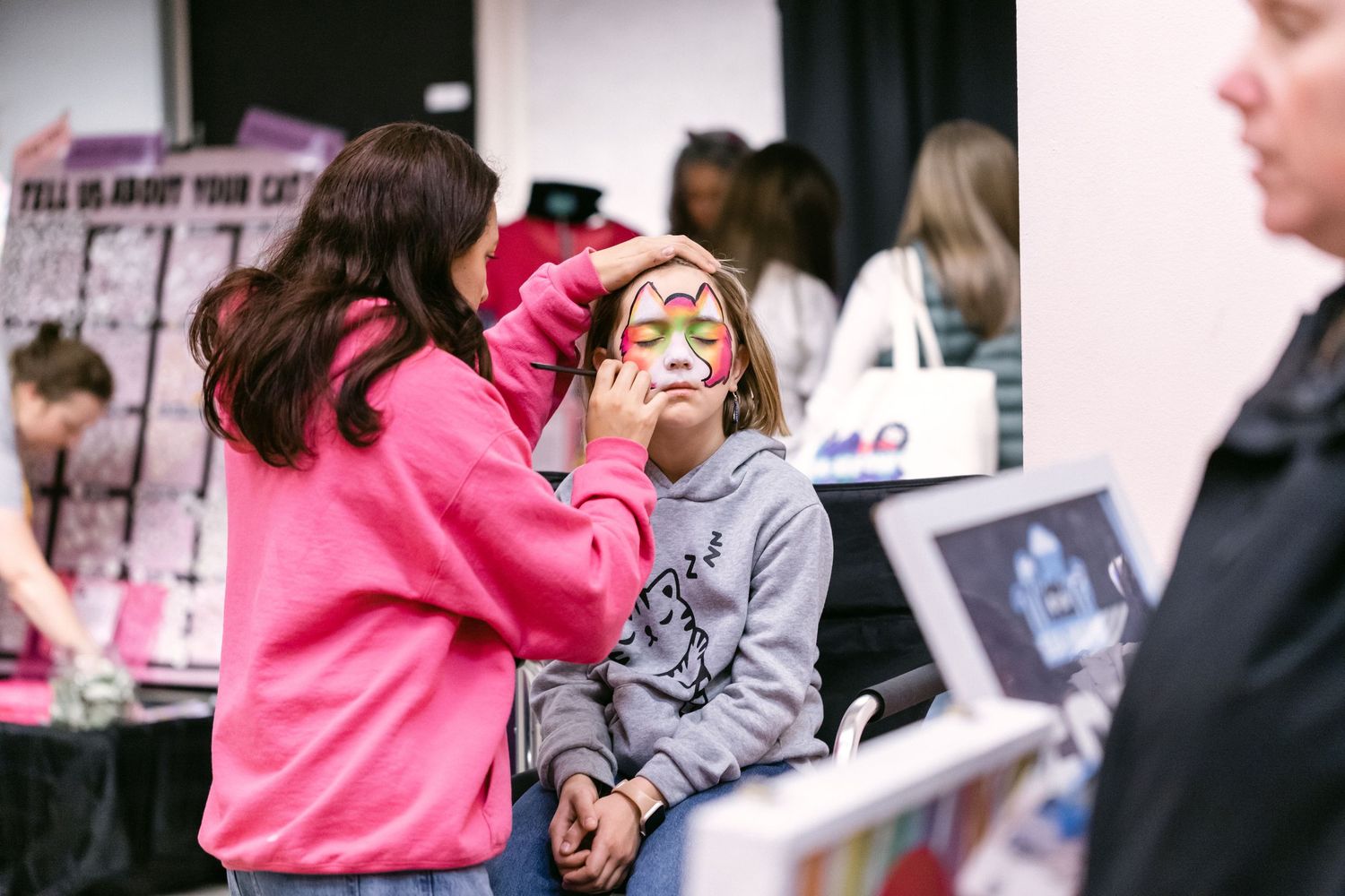 A woman in a pink hoodie applies colorful face paint on a young girl at an indoor event booth.