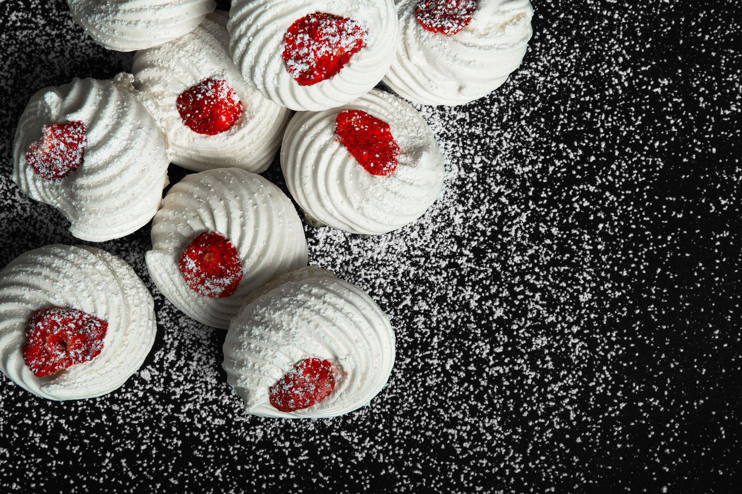 White meringue cookies topped with red raspberry jam, dusted with powdered sugar on a dark background.