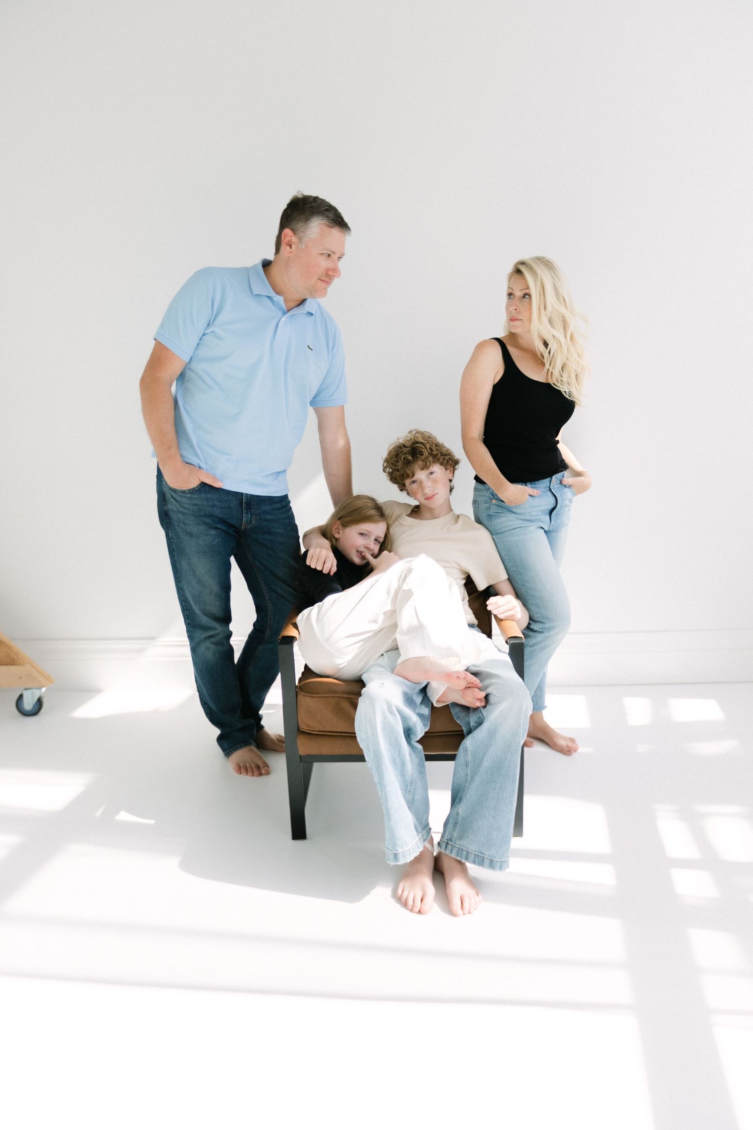 A family of four poses casually in a bright white studio with two children seated on a chair.