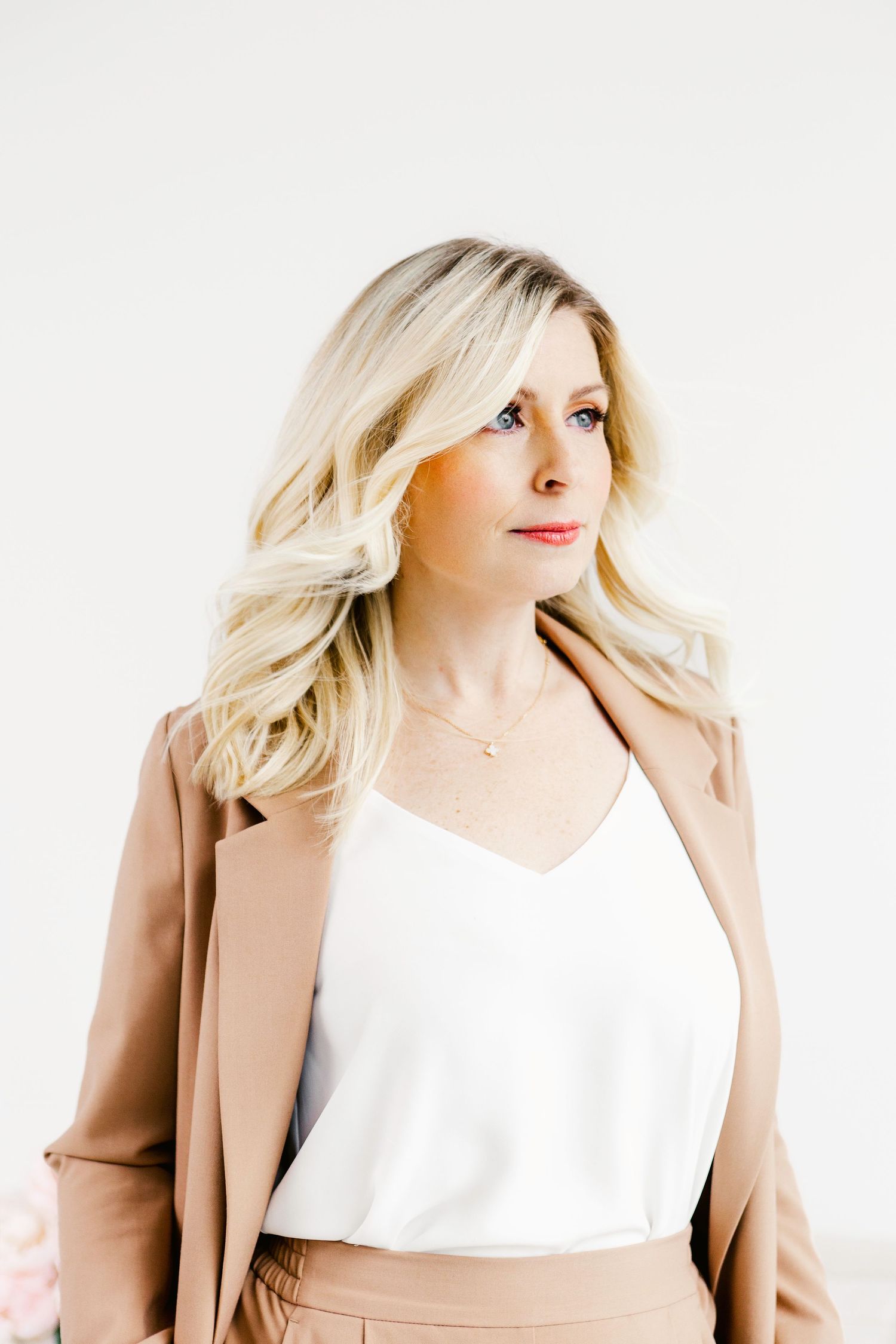 A blonde woman in a white camisole and tan blazer gazes sideways against a light background.
