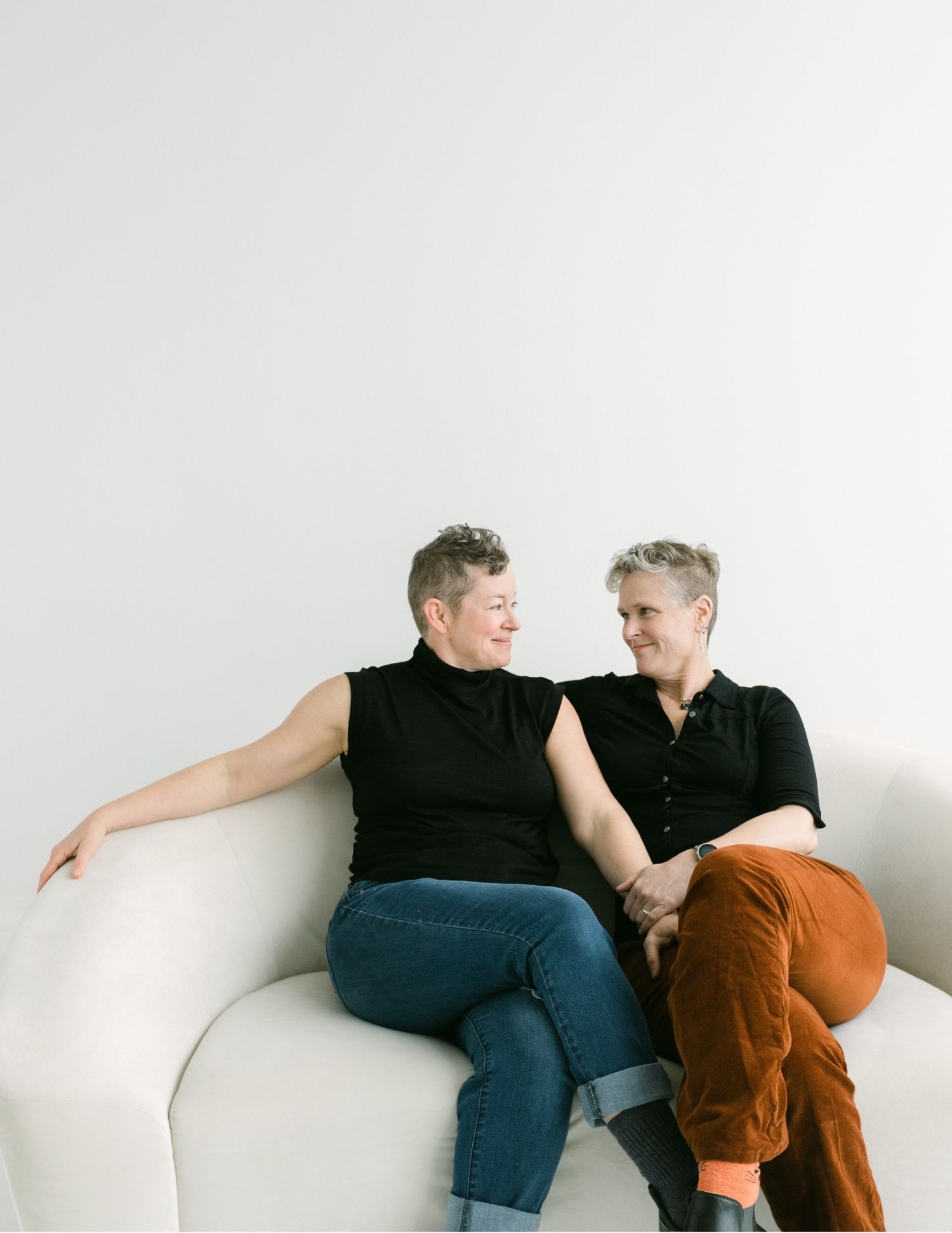 Two women sitting close on a white sofa, smiling and holding hands in a bright, minimalist setting.
