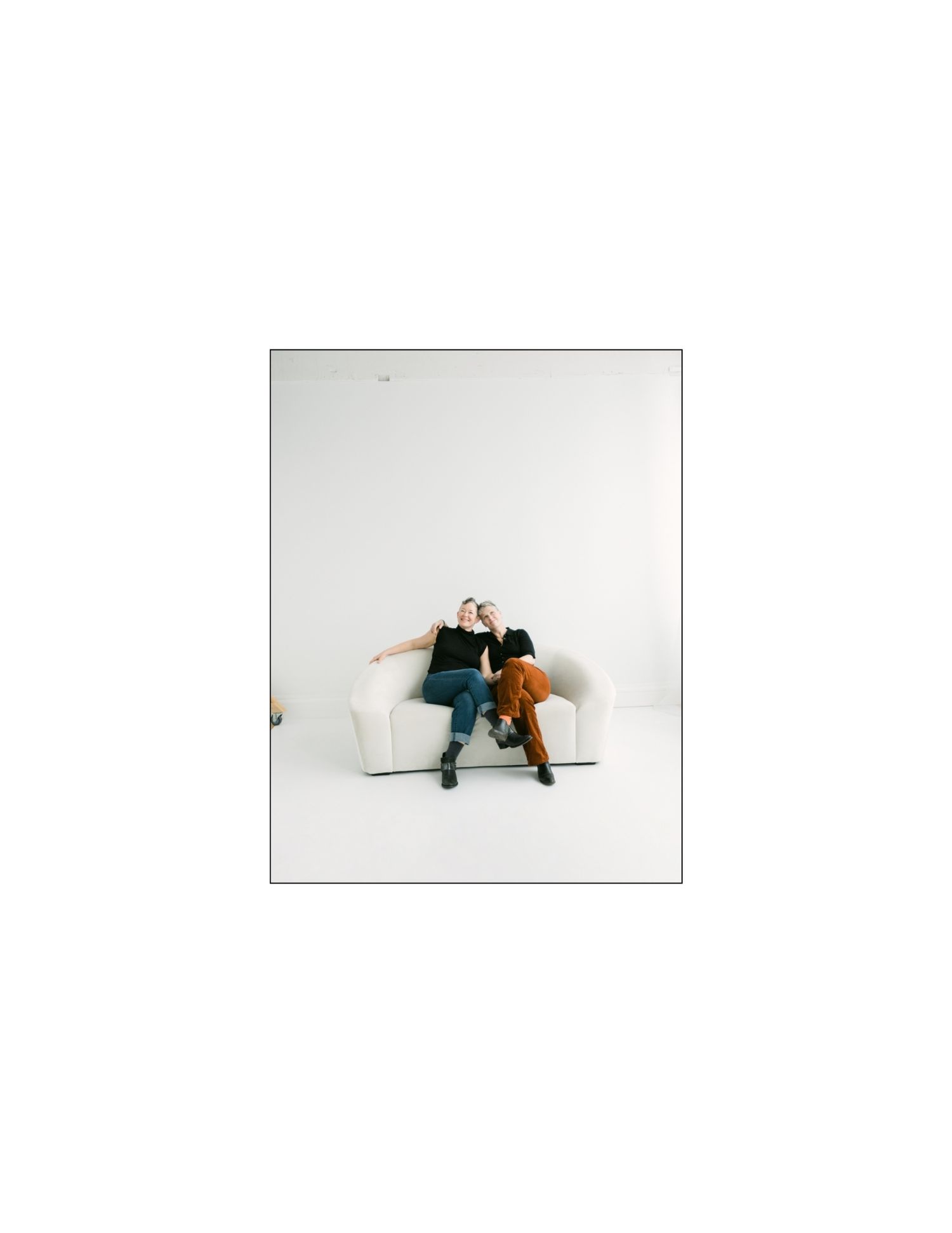 Two women sit closely together on a white couch in a minimalist white studio setting.