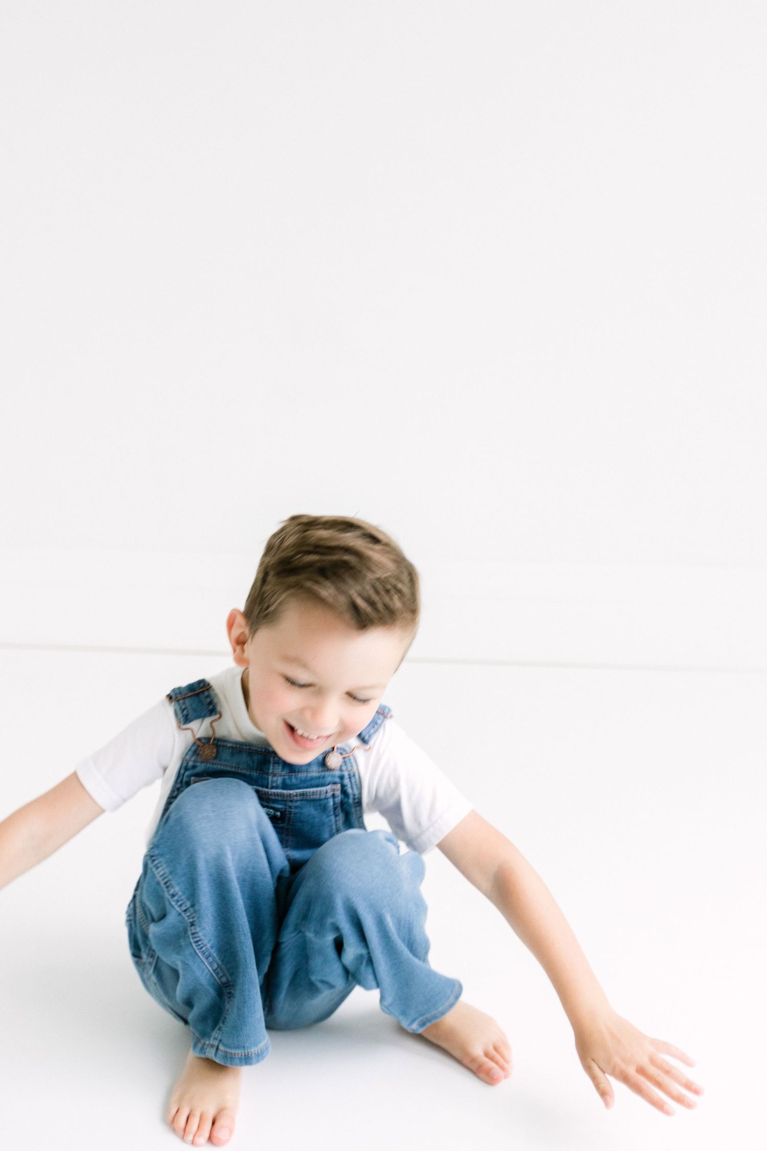 A happy young boy in denim overalls and white shirt sits on a white floor, smiling and reaching out his hand.