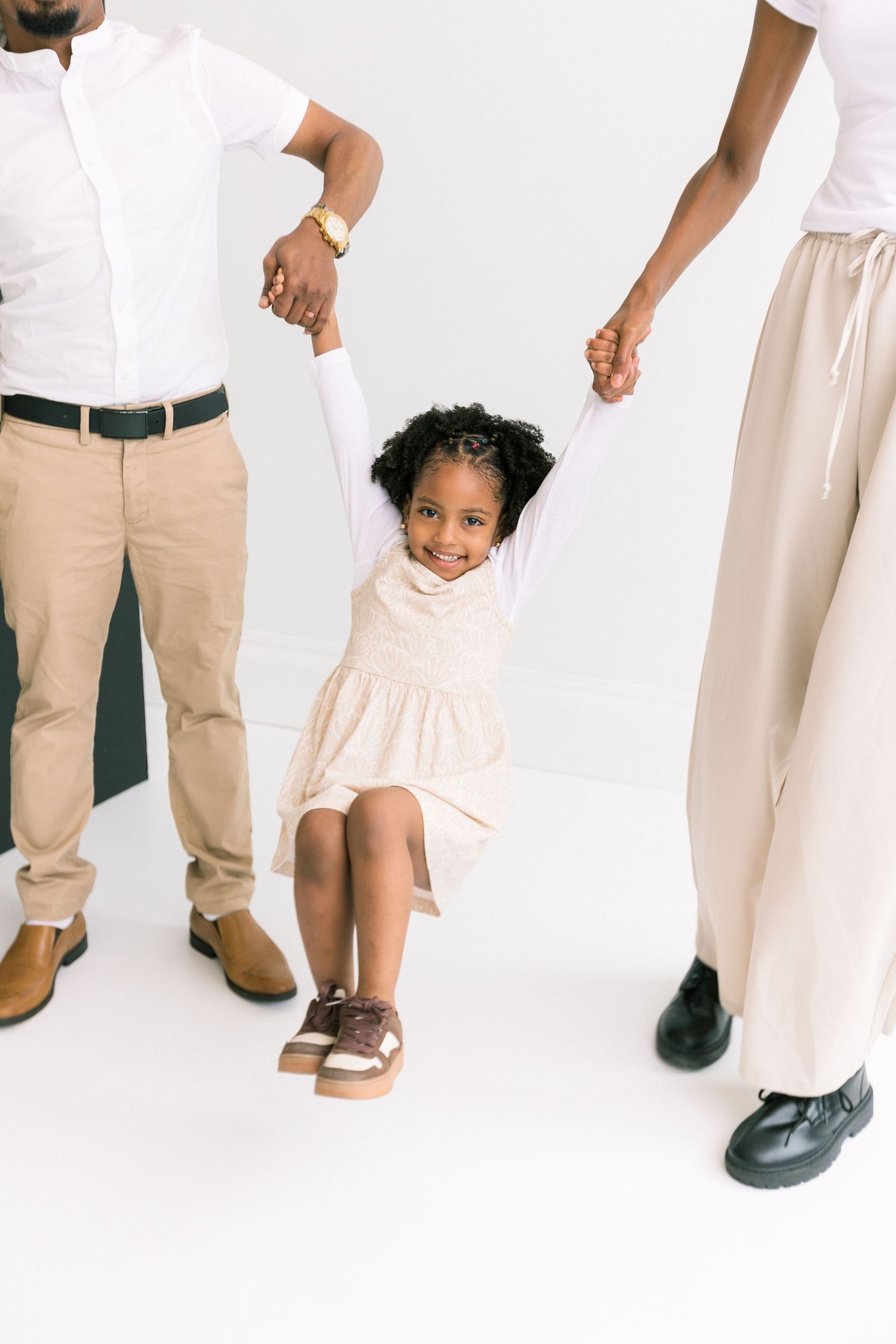 A joyful young girl in a beige dress is lifted by her parents holding her hands in a bright studio setting.