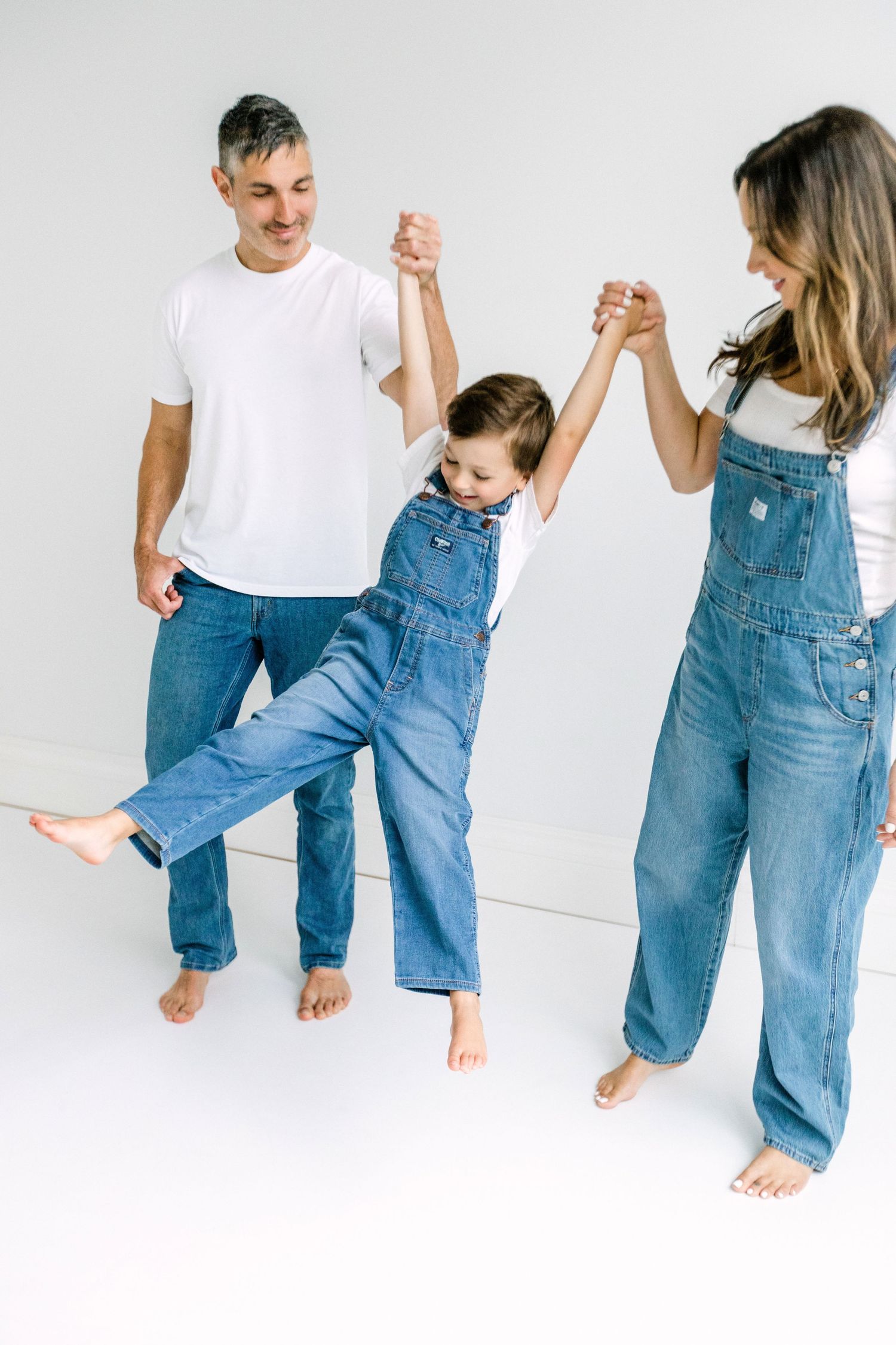 Family of three in denim overalls and jeans playfully swing child between parents against white background.