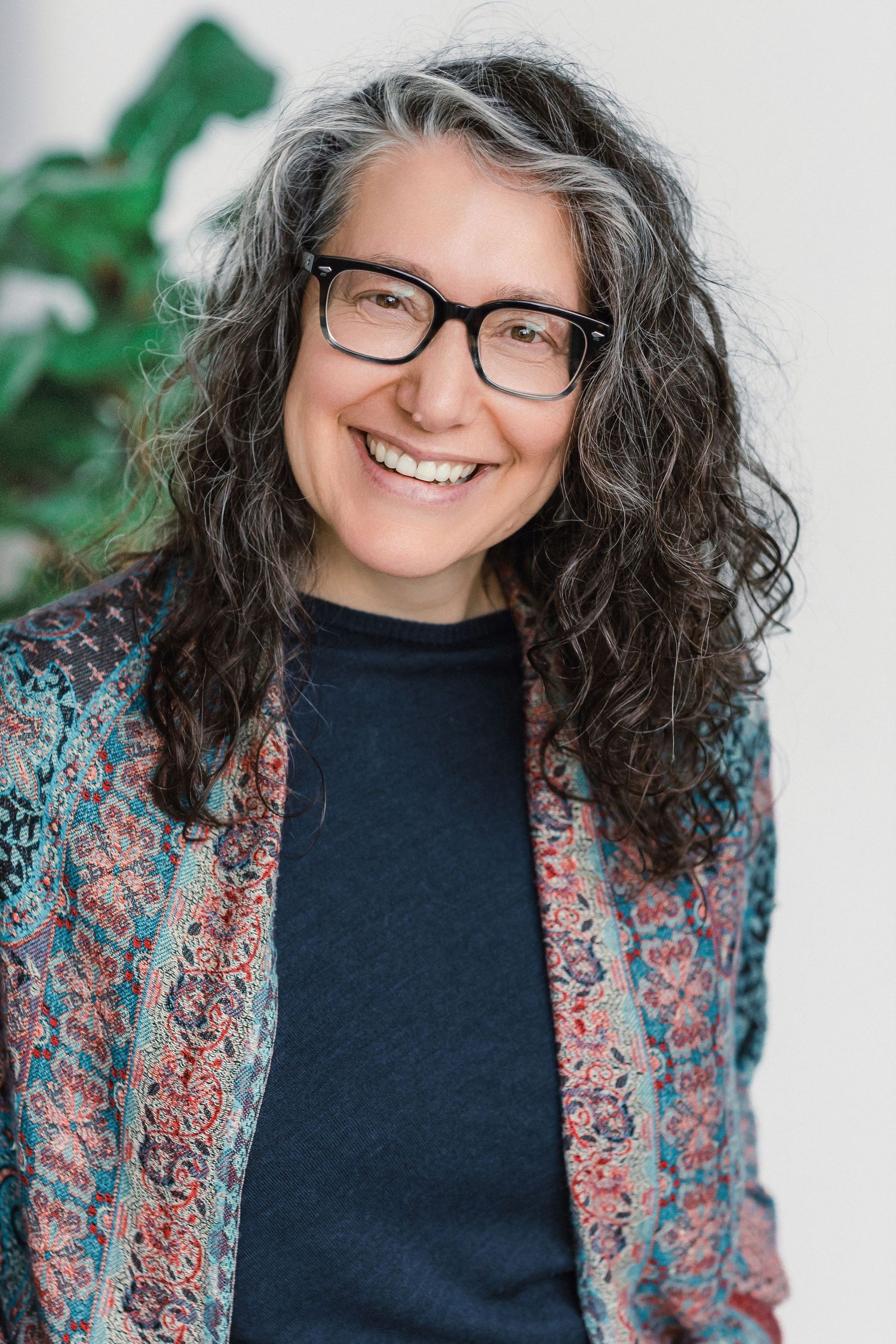 Smiling middle-aged woman with curly gray-black hair, black glasses, wearing a colorful patterned jacket over a dark shirt.