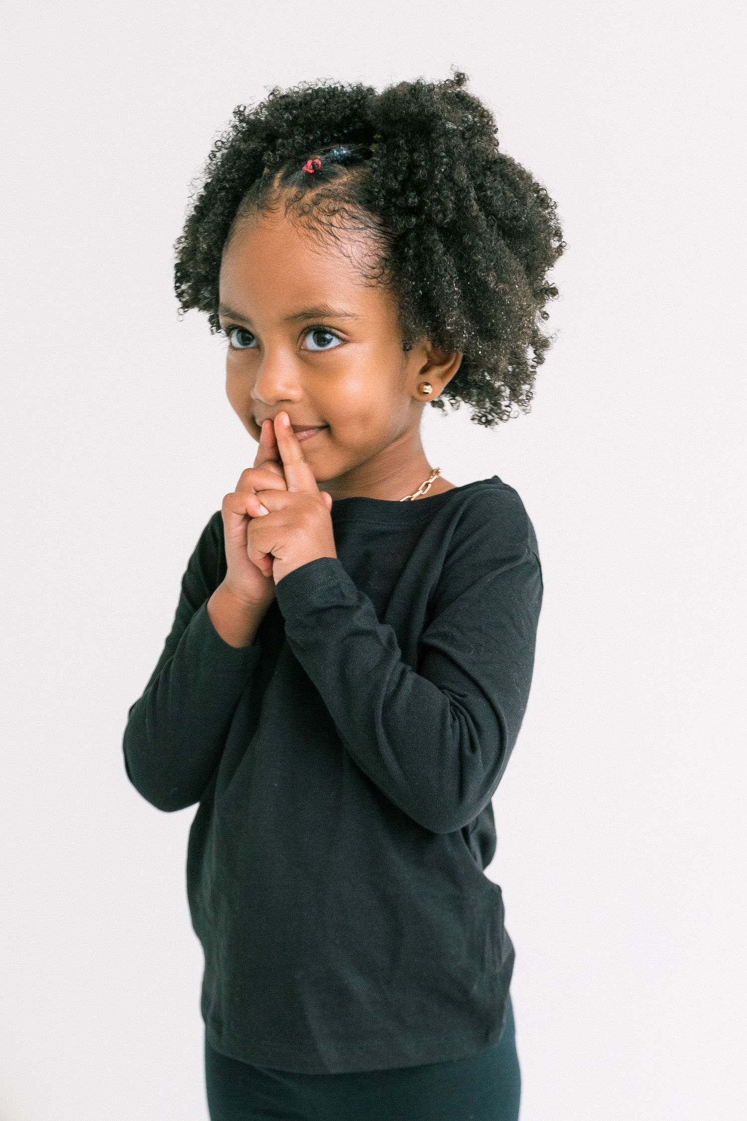 An adorable young girl with curly hair places her finger to her lips in a playful shushing gesture.