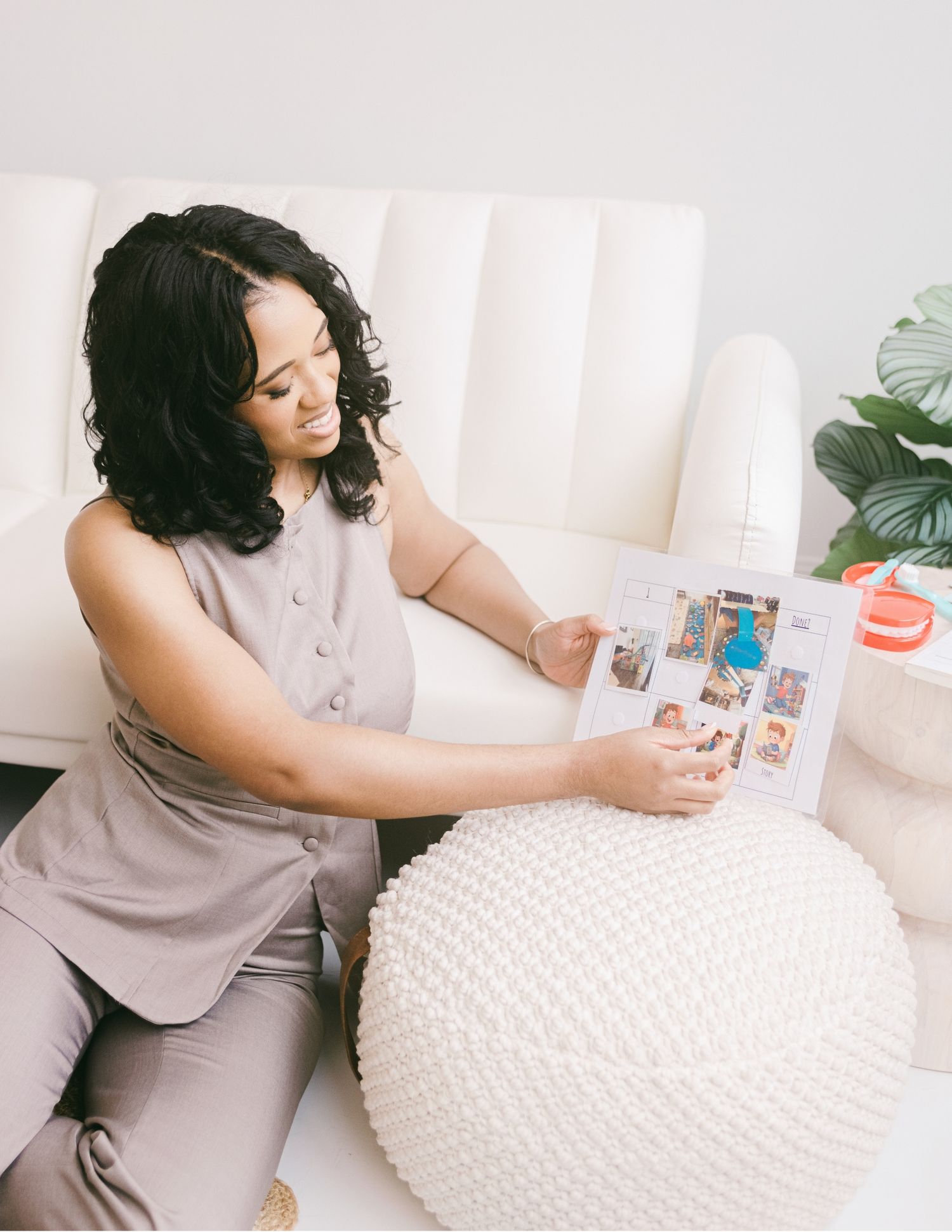 A woman in a gray outfit sits on the floor reviewing a colorful photo book near a white couch and knit pouf.