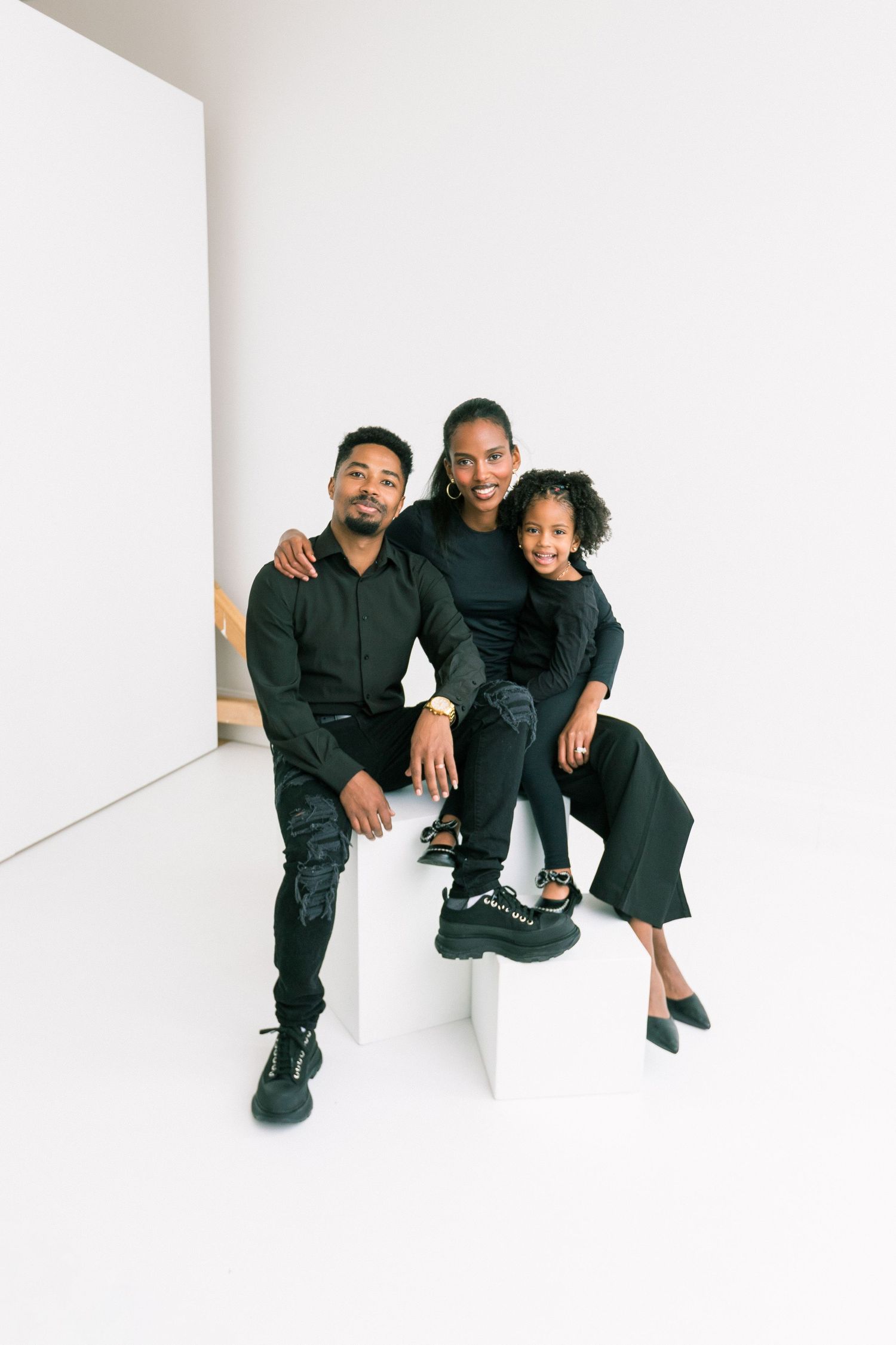 A family of three in matching black outfits pose together on a white cube in a bright studio setting.