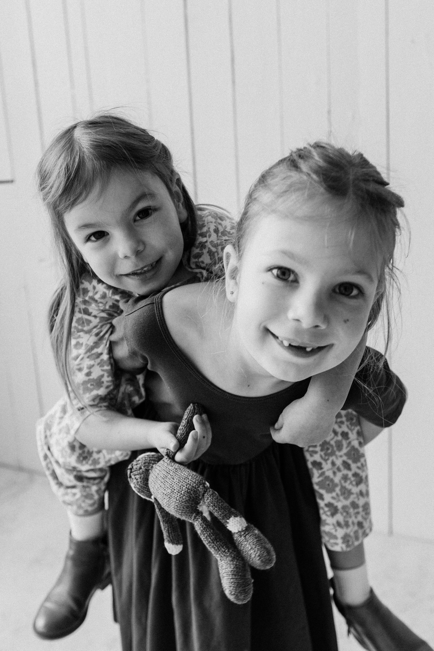 Two young sisters smile at the camera in a black and white photo, one giving the other a piggyback ride.