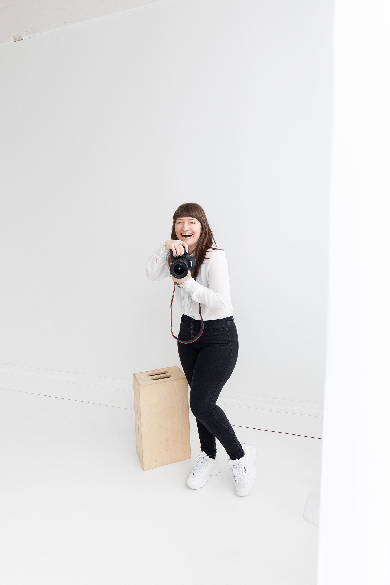 Happy female photographer laughing while holding a camera in a bright white photography studio.
