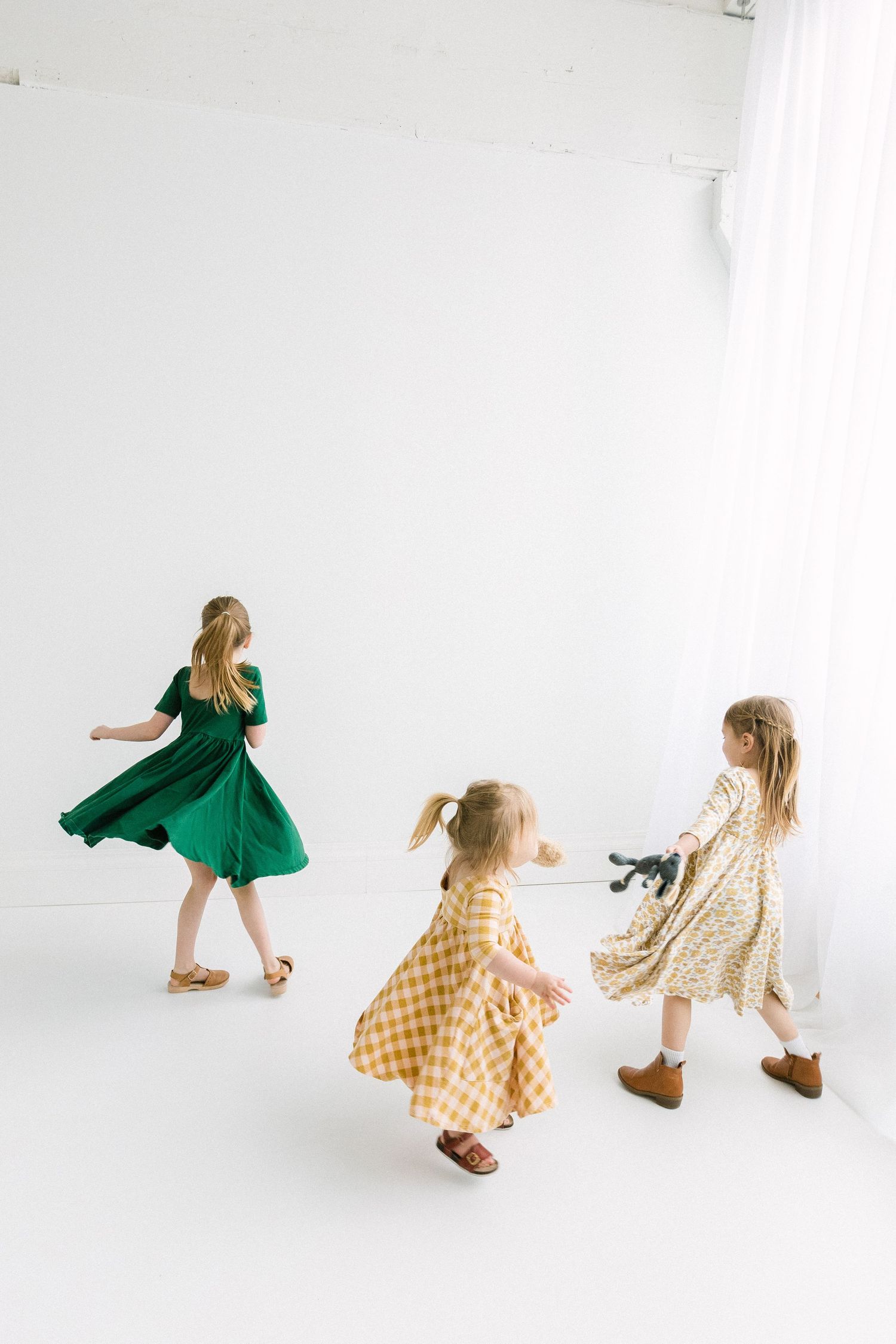 Three little girls in colorful dresses twirl and dance together in a bright white studio photography space.