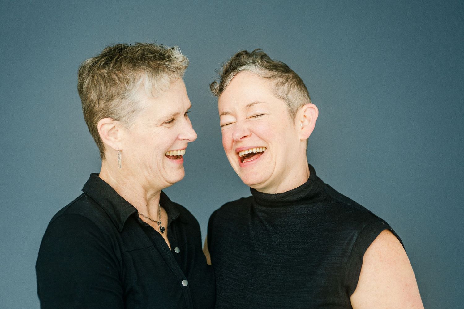 Two short-haired women in black tops laughing together against a gray studio background.