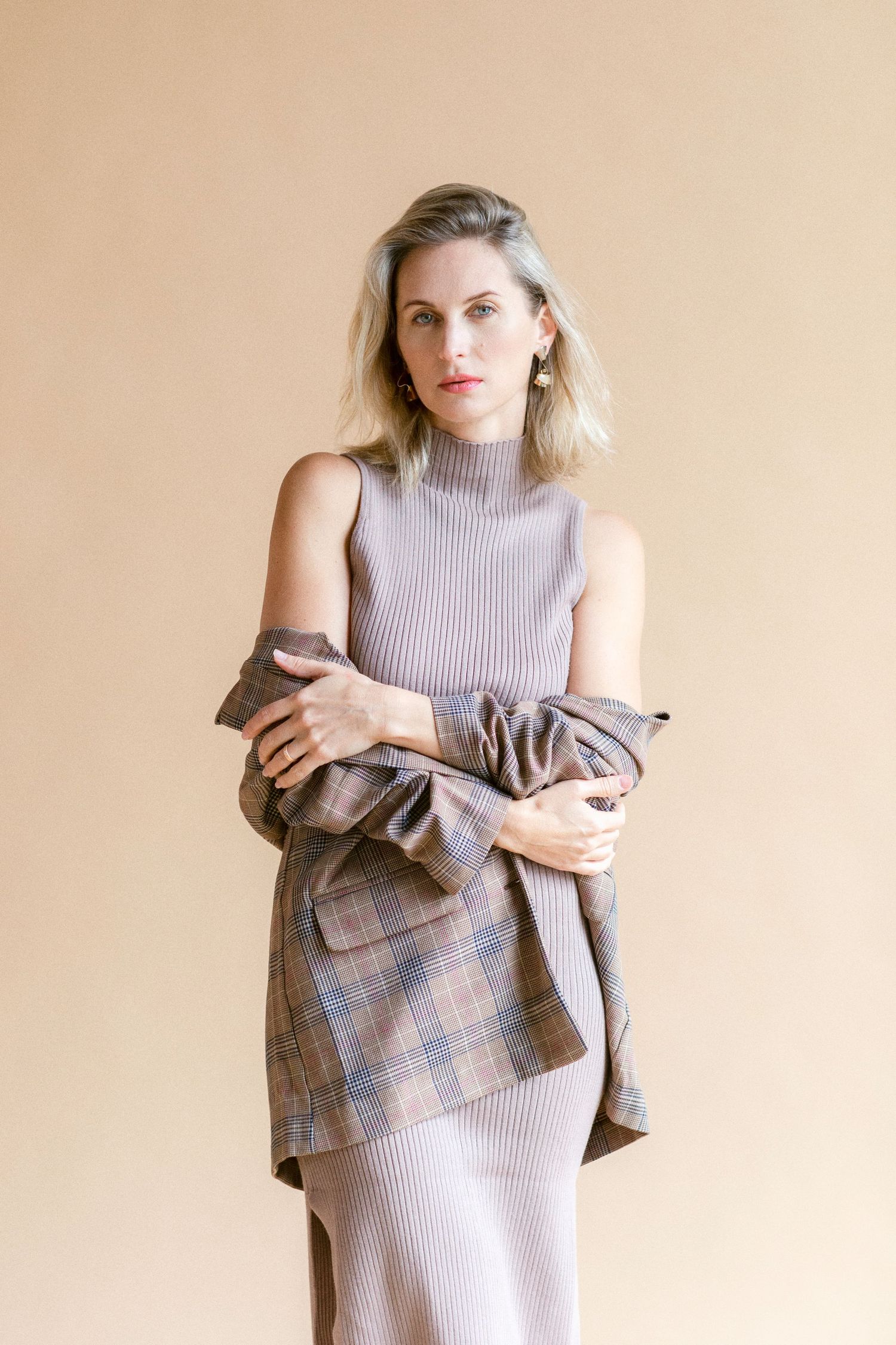 Woman in ribbed sleeveless dress with plaid blazer draped over shoulders, posing against beige background.