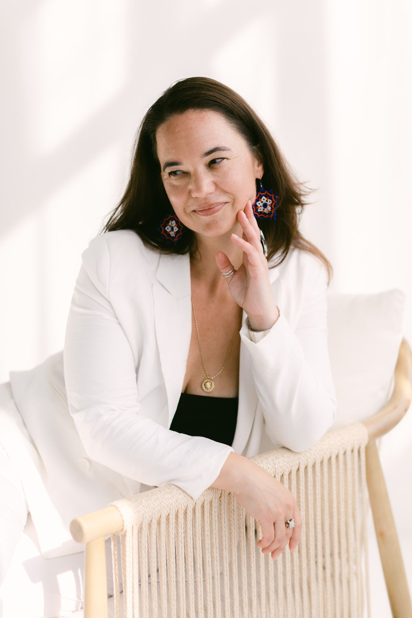 Woman in white blazer with colorful earrings sits relaxed in a wicker chair against a bright white background.