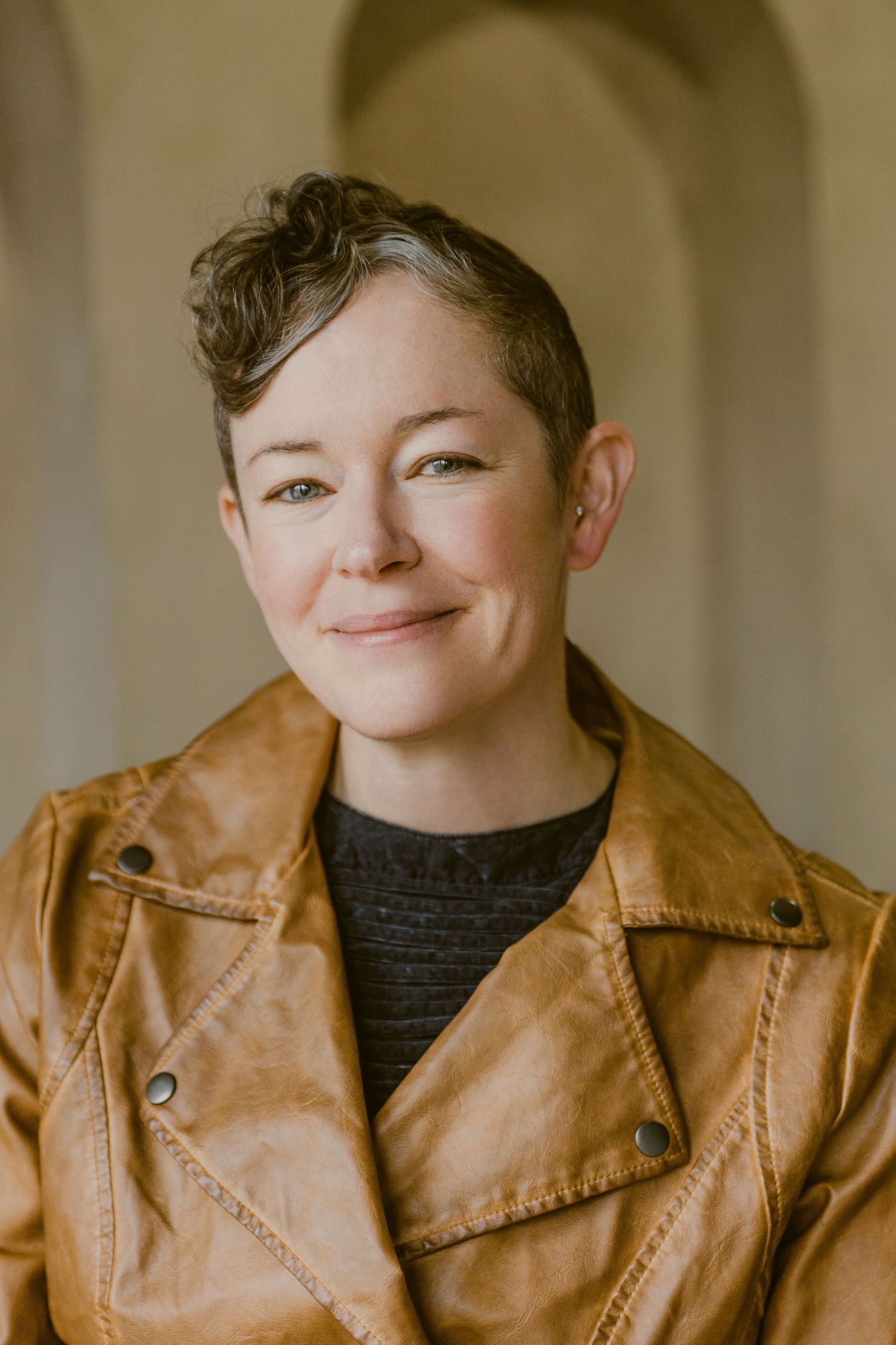A smiling person with short curly hair wearing a tan leather jacket poses for a portrait photo.