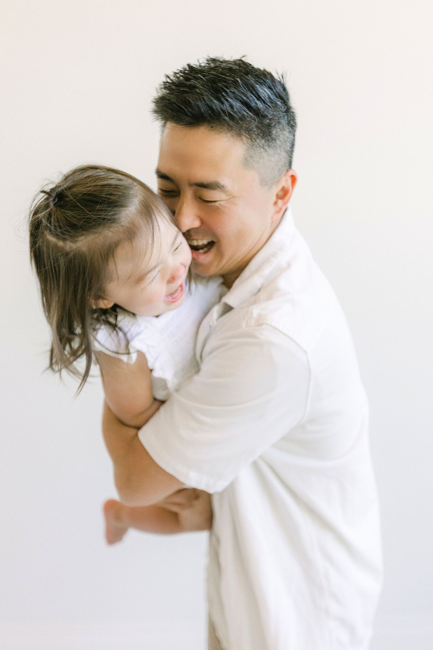 A smiling Asian man in a white shirt joyfully holds a laughing young child in a bright, minimalist setting.
