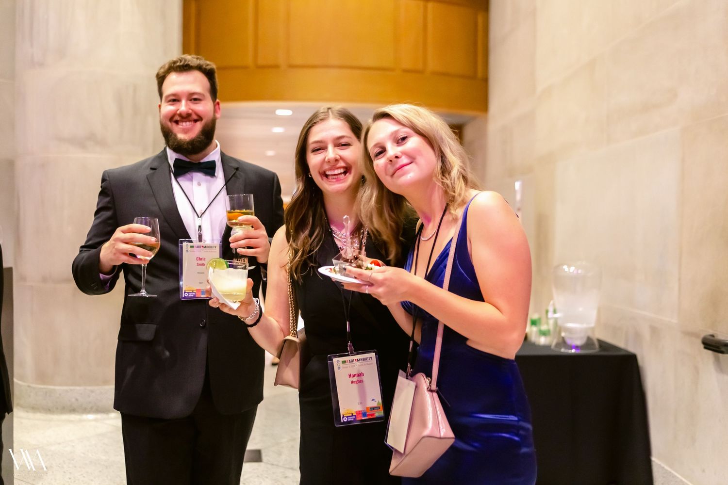 Three friends smiling and holding drinks at a formal gala event in an elegant venue.