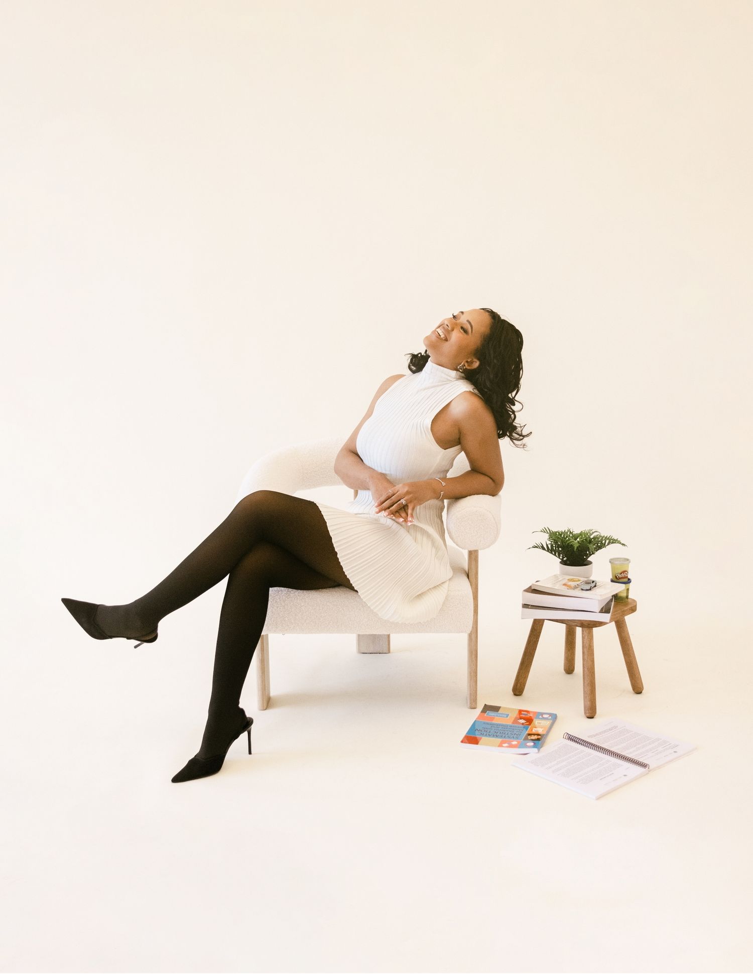 Woman in white dress and black tights sits relaxed on modern chair beside small stool with books and plant.