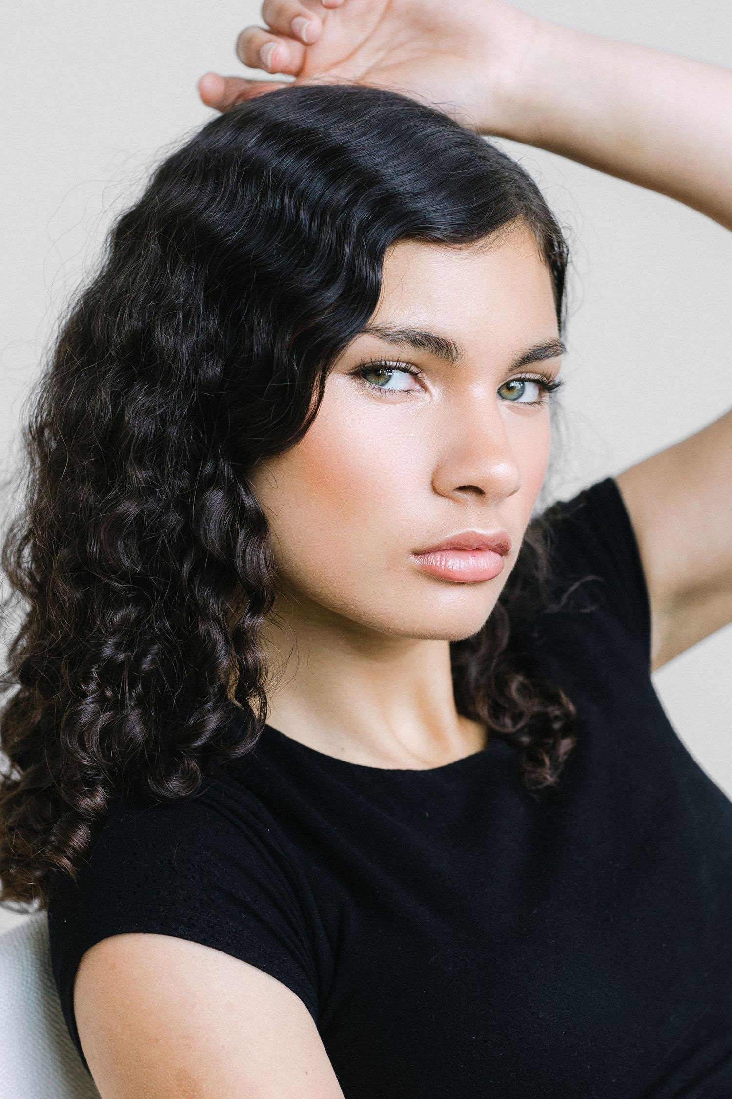 Young woman with curly dark hair and blue eyes poses in a black t-shirt against a light background.