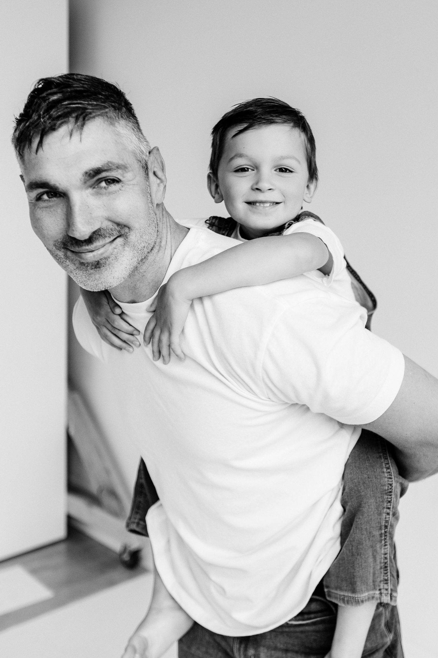 Black and white photo of a smiling man giving a happy young boy a piggyback ride indoors.