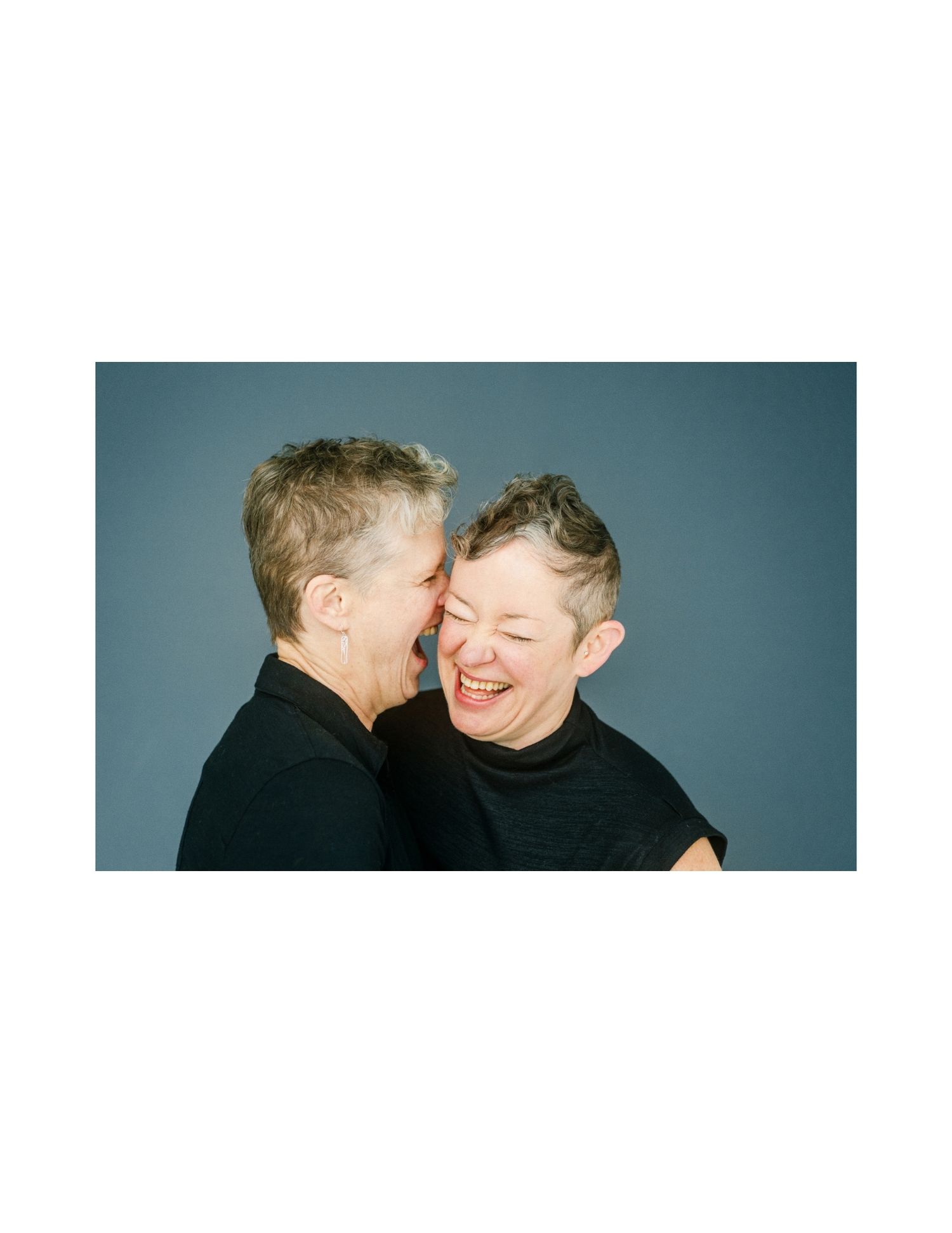 Two short-haired women in black tops share a joyful moment, one whispering while the other laughs warmly.