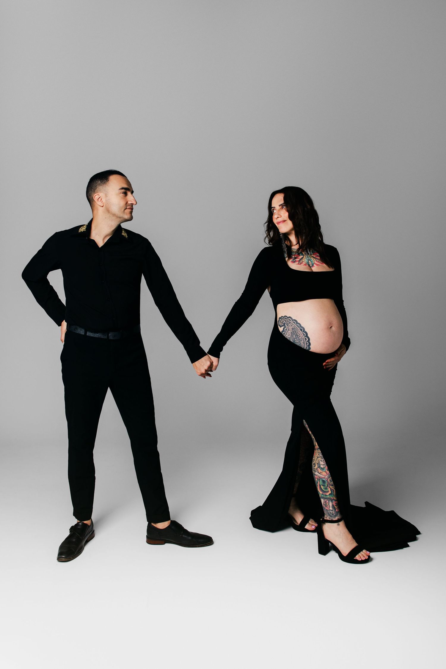 Pregnant couple in matching black outfits holding hands against gray background, woman showing baby bump with tattoos.