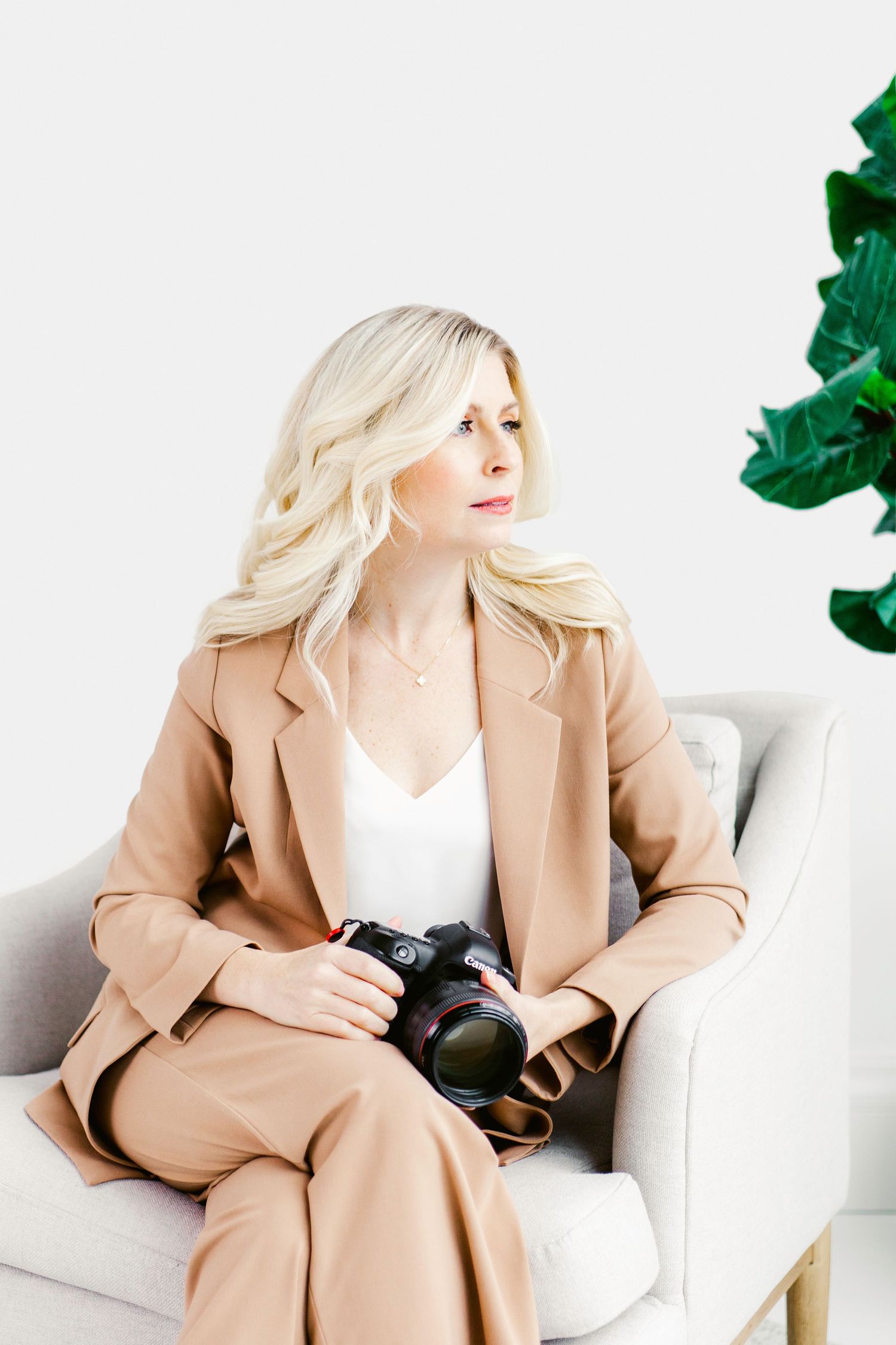 Blonde woman in tan blazer sitting in armchair holding a camera, professional photographer portrait with plant.