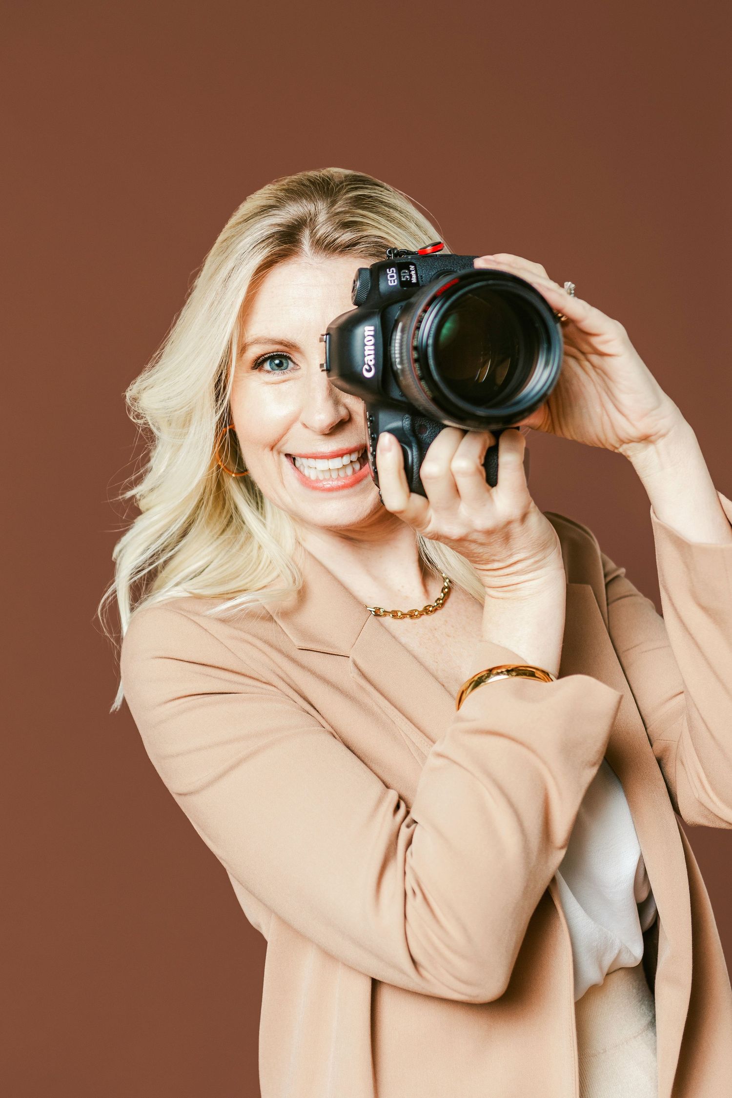 Smiling blonde woman holding a Canon DSLR camera up to her face, wearing a beige blazer against a brown background.