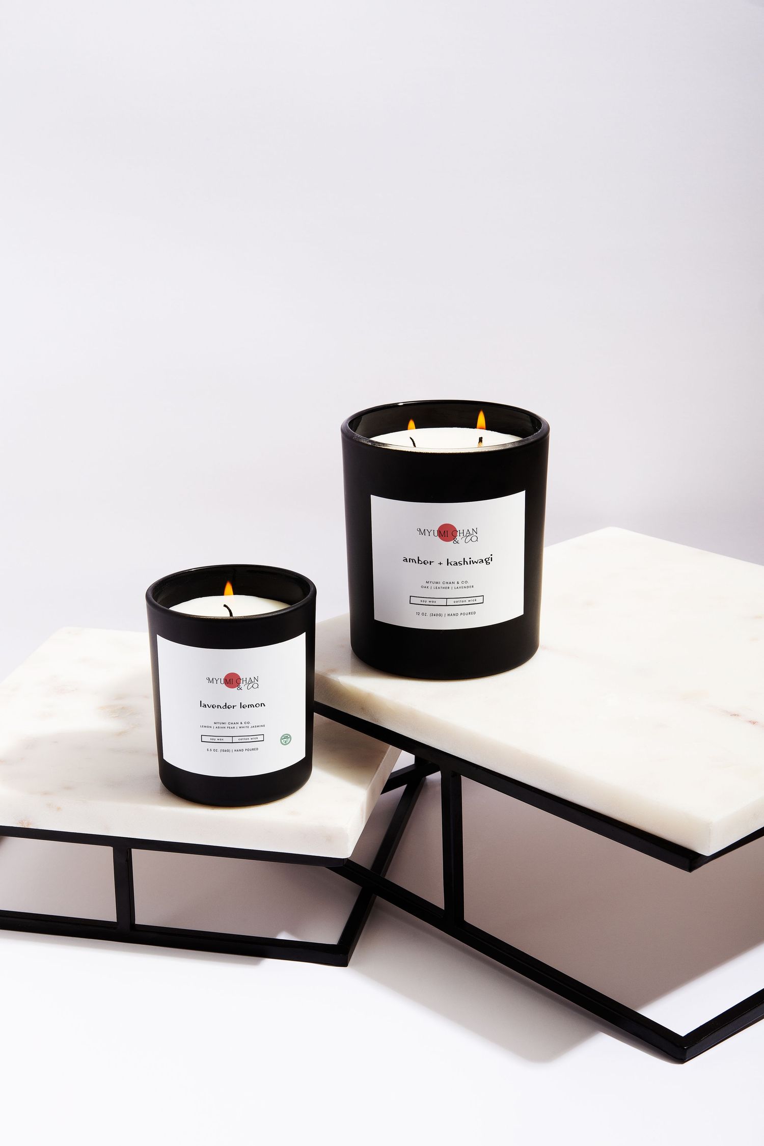 Two lit black glass scented candles in amber and lavender lemon fragrances displayed on marble and metal surfaces.