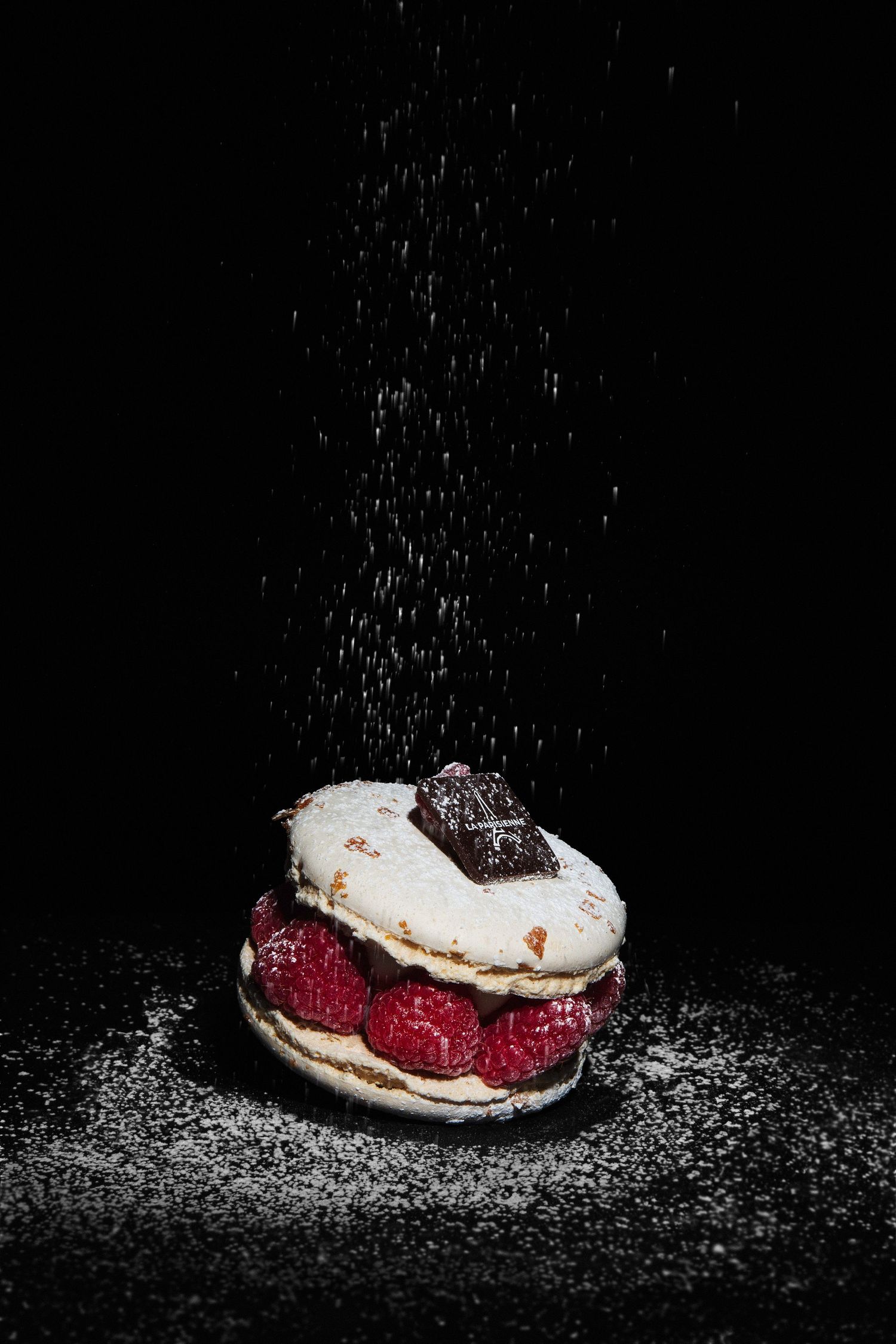 Elegant raspberry macaron dessert dusted with powdered sugar falling from above, topped with chocolate on black background.