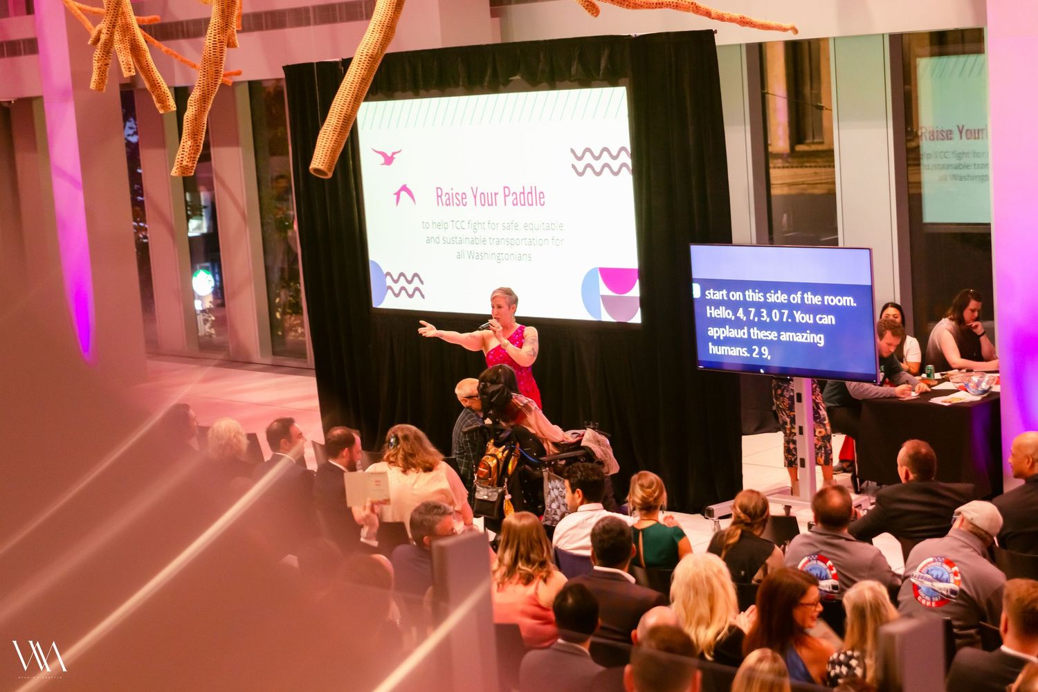Blonde presenter speaks on stage with pink lighting to a seated audience at a formal event.
